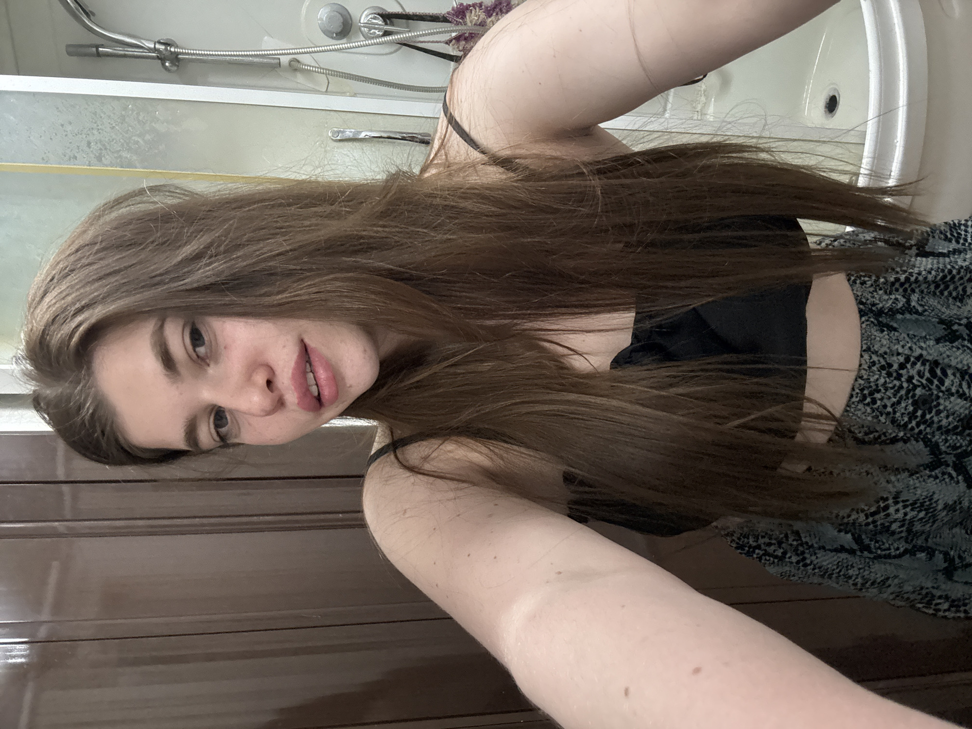 A young woman with long brown hair is taking a selfie indoors. She is wearing a black top with a spaghetti strap. Part of a bathroom is visible, with a shower and sink. The lower portion of a wooden door is also visible. The location is Мещёры, Russia.