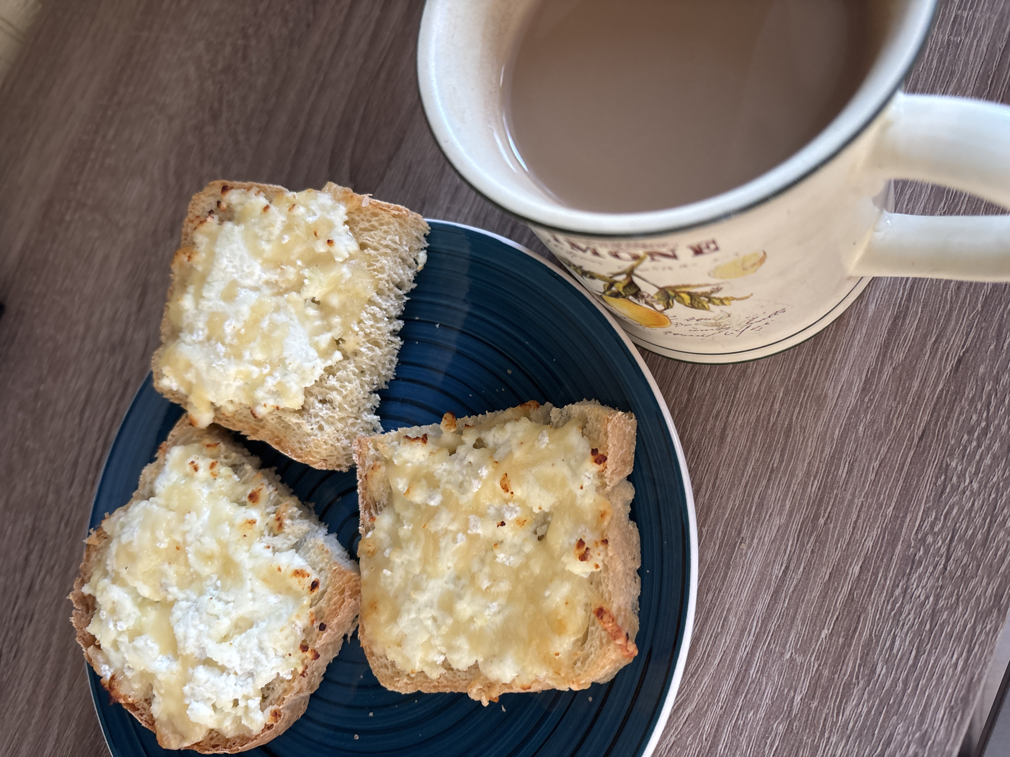 Three slices of toasted bread topped with a white cheese spread are arranged on a blue plate. A mug filled with a light brown liquid, possibly tea or coffee, sits to the right of the plate. The items are placed on a wooden surface with a visible grain pattern. The scene appears to be indoors, likely in a kitchen or dining area. There is no indication of the specific location within Naberezhnye Chelny, Russia.