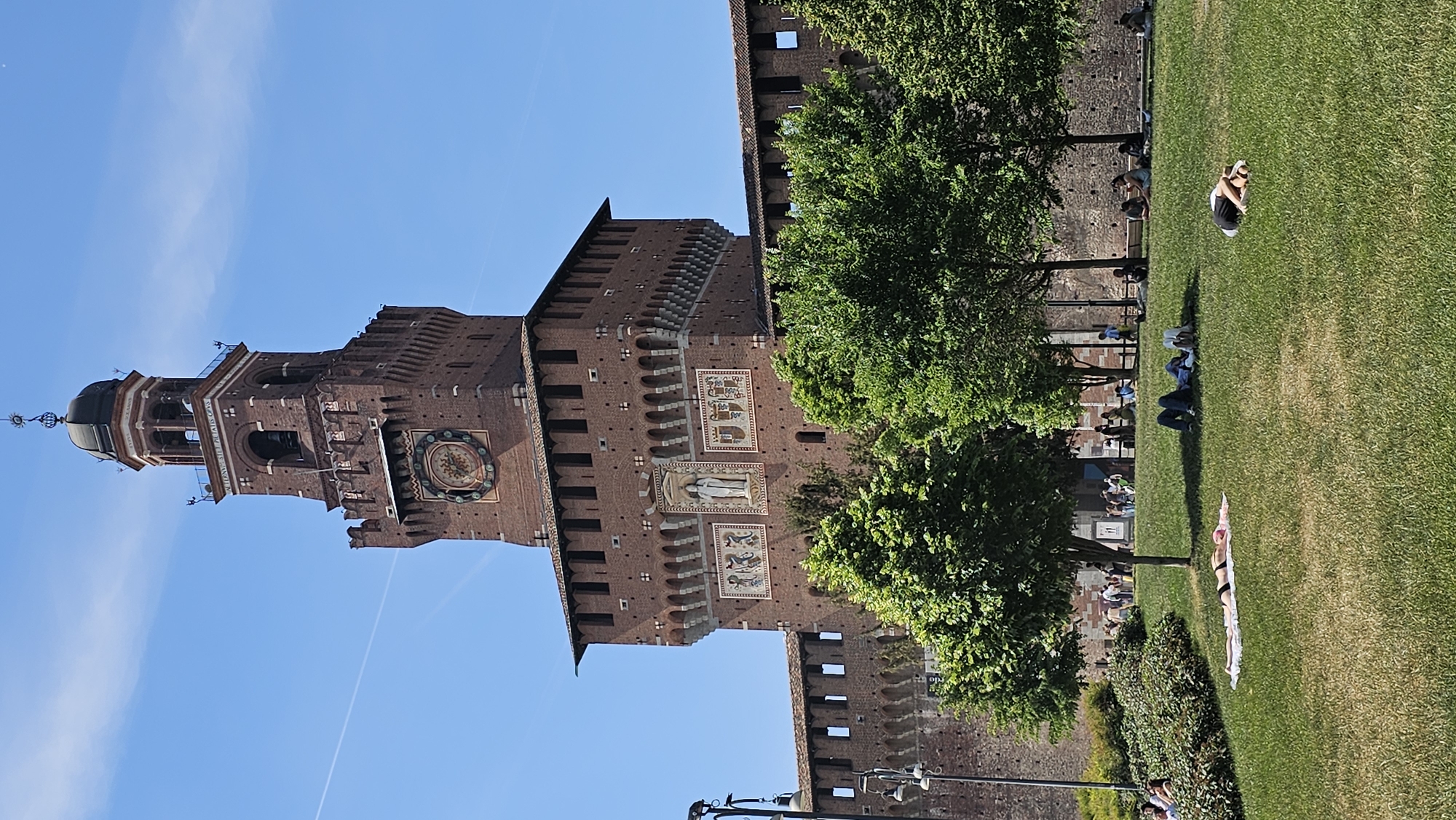 The image depicts the Sforza Castle in Milan, Italy. The castle, constructed of red brick, features a prominent clock tower, battlements, and decorative panels. A green lawn extends alongside the castle's base, where several people are relaxing, sunbathing, and sitting under trees. The sky is blue with a few streaks of clouds.