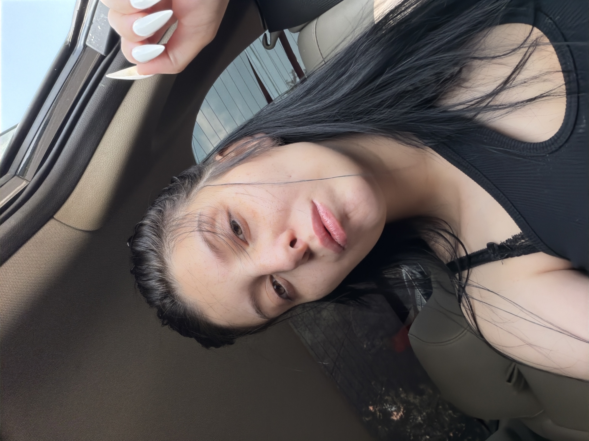 A young woman with long, dark hair and fair skin is taking a selfie in a car. She is wearing a black, ribbed tank top with a cutout detail. Her nails are long and painted white. The interior of the car is visible, with beige seats and a dark headliner. The lighting suggests it is daytime, with sunlight filtering in through the windows. The background outside the car window shows trees and a cloudy sky. There is no discernible location in the city of Tbilisi, Georgia, indicated by the visual information in the image.