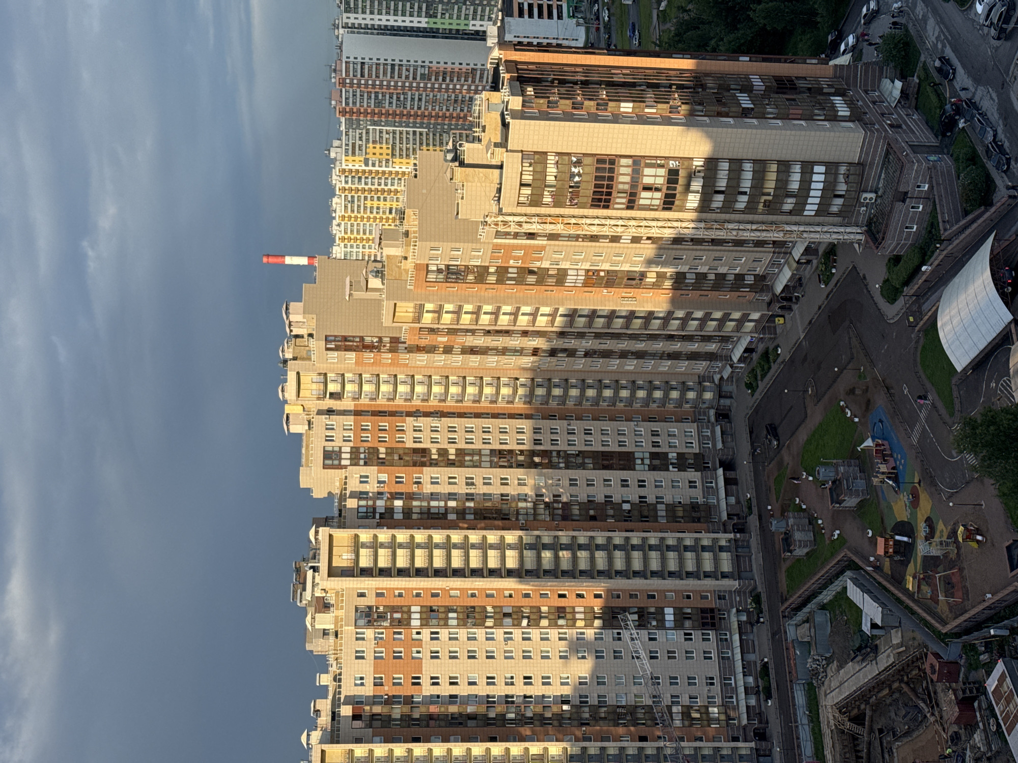 The image captures an aerial view of a residential complex in Saint Petersburg, Russia, during what appears to be late afternoon or early evening, as evidenced by the long shadows cast by the buildings. The primary focus is on several tall, multi-story apartment buildings with a mixture of beige, orange, and gray facades. Many windows and balconies are visible, and some apartments show lights on.
In the foreground, there is a construction site with scaffolding, earthworks, and materials. Adjacent to the construction area is a playground with colorful equipment, including slides and swings, and a blacktop surface with painted markings.
Several vehicles are parked on the streets surrounding the buildings. A few individuals can be seen walking on the sidewalks and in the playground area. The sky above is a mix of dark, stormy clouds and patches of brighter sky, suggesting a transition in weather. A tall, red and white striped chimney is visible in the distance, rising above the rooftops of other buildings. The overall scene depicts a typical urban residential environment with ongoing development and community spaces.
