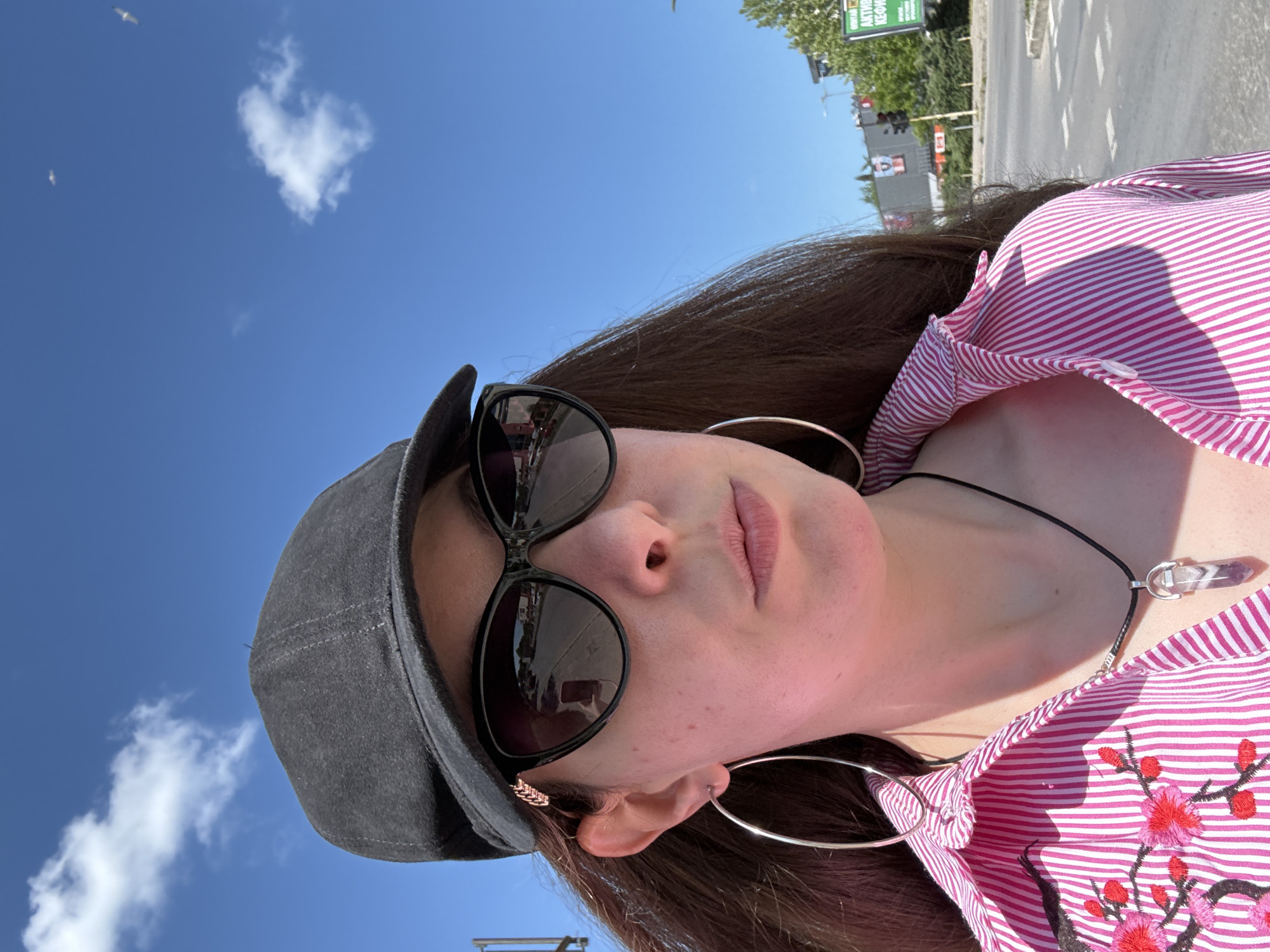 A female subject in Unknown, Unknown, is pictured in a medium shot selfie taken outdoors during daylight. The environment consists of a partly cloudy blue sky, suggesting a sunny day. In the background, a street scene is visible with traffic lights, pedestrian crossing, and a green signage board, which partially reads "AKТИВЕН КЕОДОП".
The subject is wearing a gray cap and large, round sunglasses, which reflect the surrounding scene. Her outfit consists of a pink and white striped shirt adorned with embroidered red and black floral patterns on the lower right side. She wears large, hoop earrings and a necklace with a crystal pendant. The focus of the image is on her face and upper body, capturing a direct, yet casual, presentation of her appearance in an urban environment.