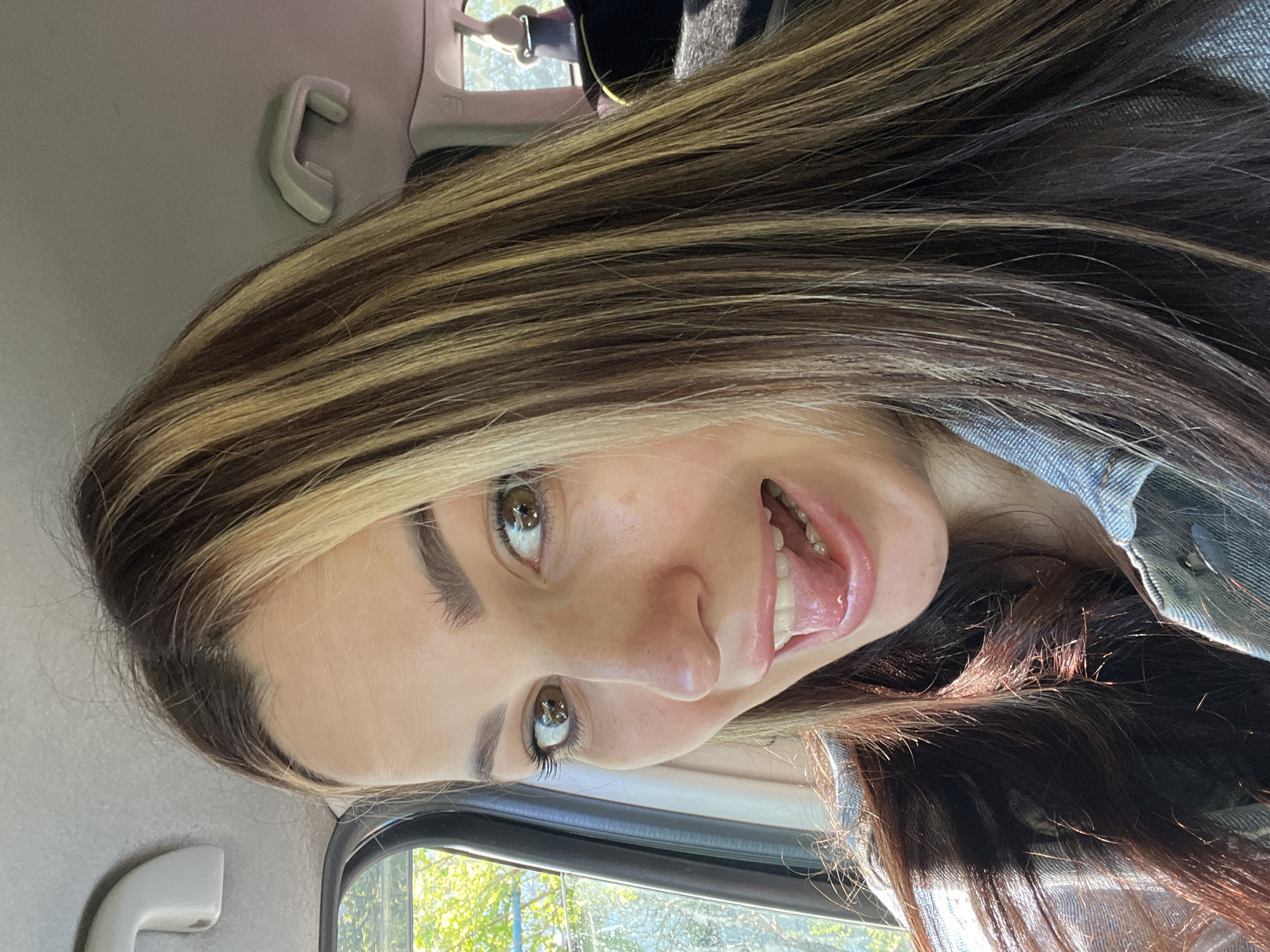 A young woman with brown eyes and highlighted brown and blonde hair is in a car, possibly a taxi, looking at the camera. Her mouth is open, and her tongue is slightly extended. She appears to be wearing a denim jacket. The interior of the car includes a window, a headliner grab handle, and a sun visor attachment. Outside the window, greenery suggests a tree-lined street. The location is Unknown, Unknown.