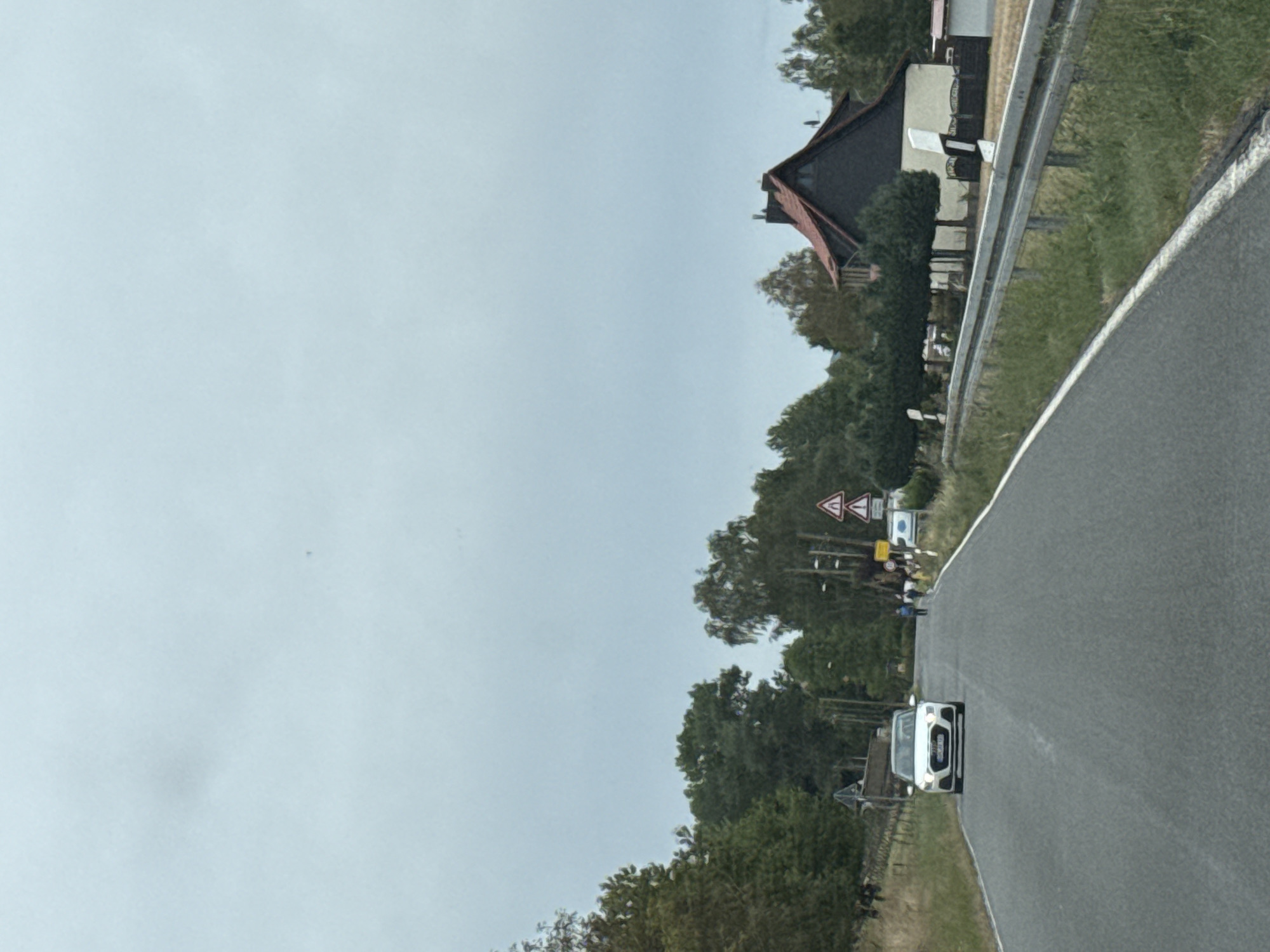 The image shows a view from a vehicle traveling on a road in or near Ellrich, Germany. The road curves slightly to the right, and a white Audi is visible in the lane ahead, stopped or moving slowly. To the right of the road, there is a metal guardrail bordering a grassy area. Beyond the guardrail, a house with a dark, steep-pitched roof and a light-colored facade is visible, surrounded by trees. Several traffic signs are present along the roadside, including triangular warning signs. In the distance, a few people are walking on the side of the road. The sky is overcast, suggesting a cloudy day. The overall environment appears to be rural or suburban.