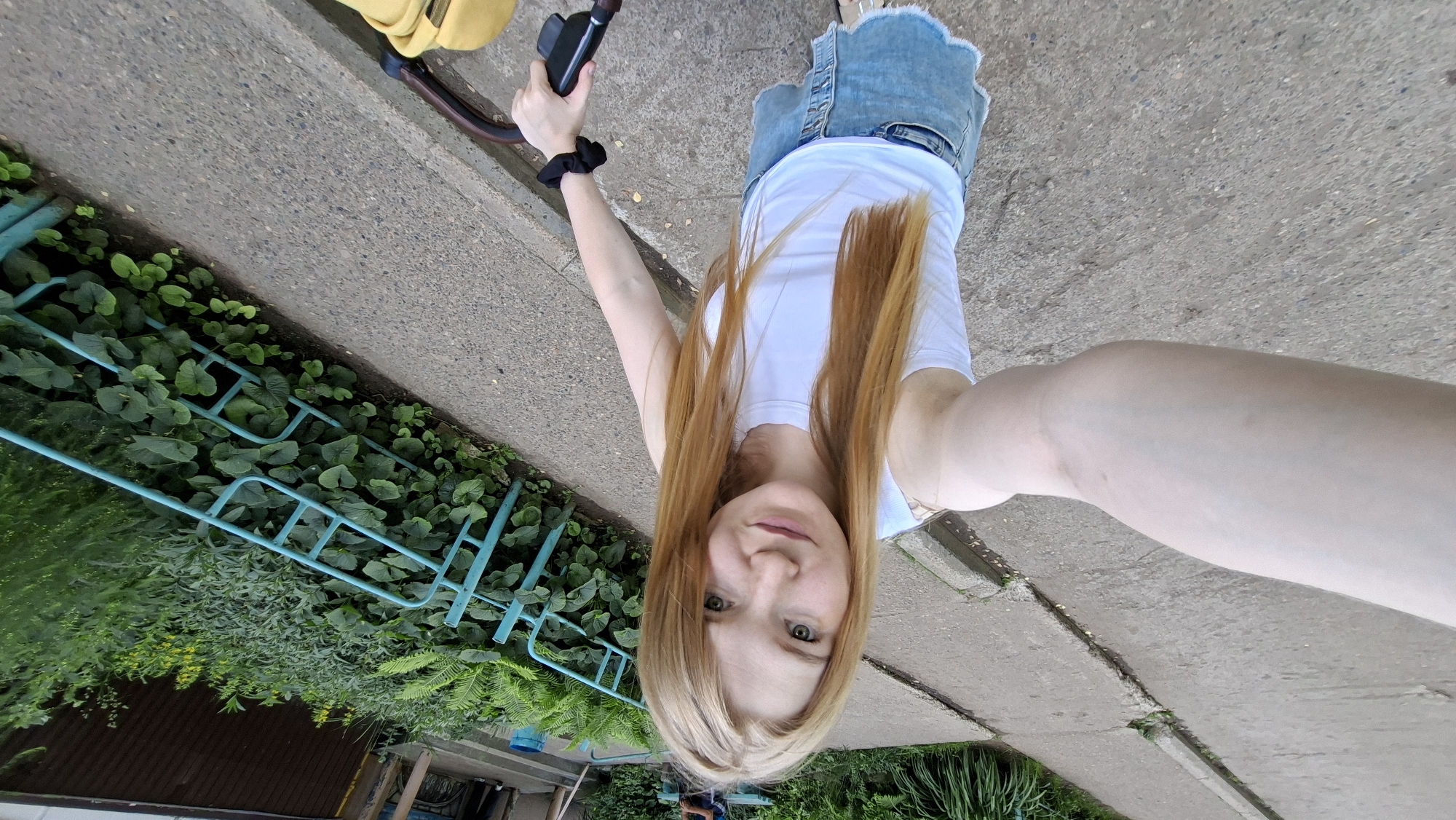 The image shows a young woman with long, reddish-blonde hair taking a selfie outdoors. She is wearing a white tank top and a denim skirt. She is holding a dark-colored object in her right hand, possibly a camera or a phone. Her left arm is bent at the elbow, and she appears to be standing or walking on a paved surface.
The background includes lush green foliage and a light blue metal fence. The ground is made of concrete slabs with some cracks and weeds growing through them. A portion of a yellow backpack is visible at the top of the image. The lighting suggests it is daytime, likely with overcast or diffused sunlight given the even illumination. The overall setting appears to be a park or a residential area with greenery. There are no visible signs or text in the image. The woman's expression is neutral, and the photo has a casual, candid feel. The location context provided indicates Chaikovsky, Russia, which is consistent with the outdoor, natural setting.