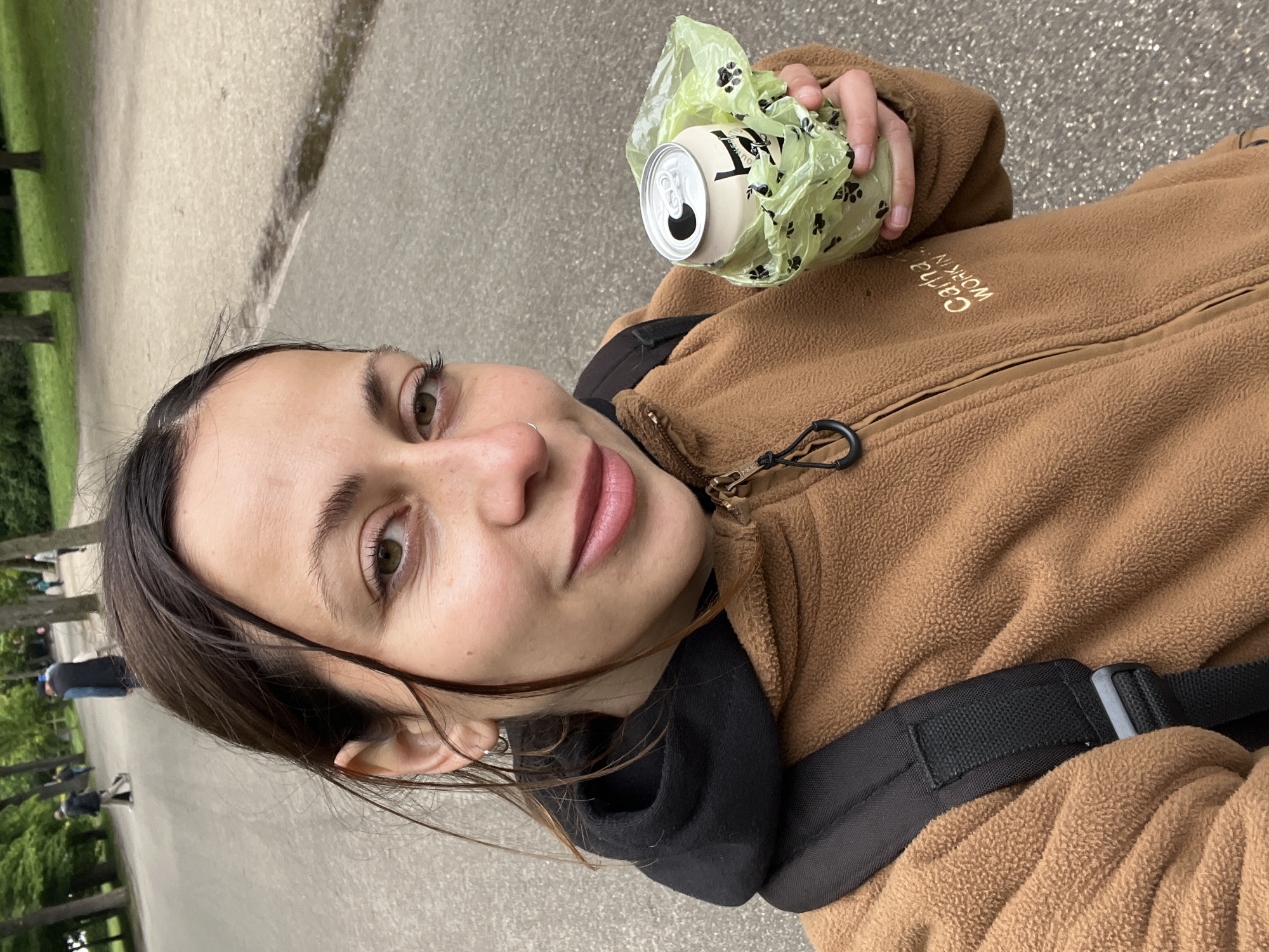 The image is a selfie of a woman holding a can of drink in a light green plastic bag with black paw print designs. She is wearing a brown fleece jacket over a black hooded top, and a black bag strap is visible. The background shows a gray paved path with trees and grass on the left side. People are faintly visible in the background. This is in a park in Amsterdam, Netherlands.