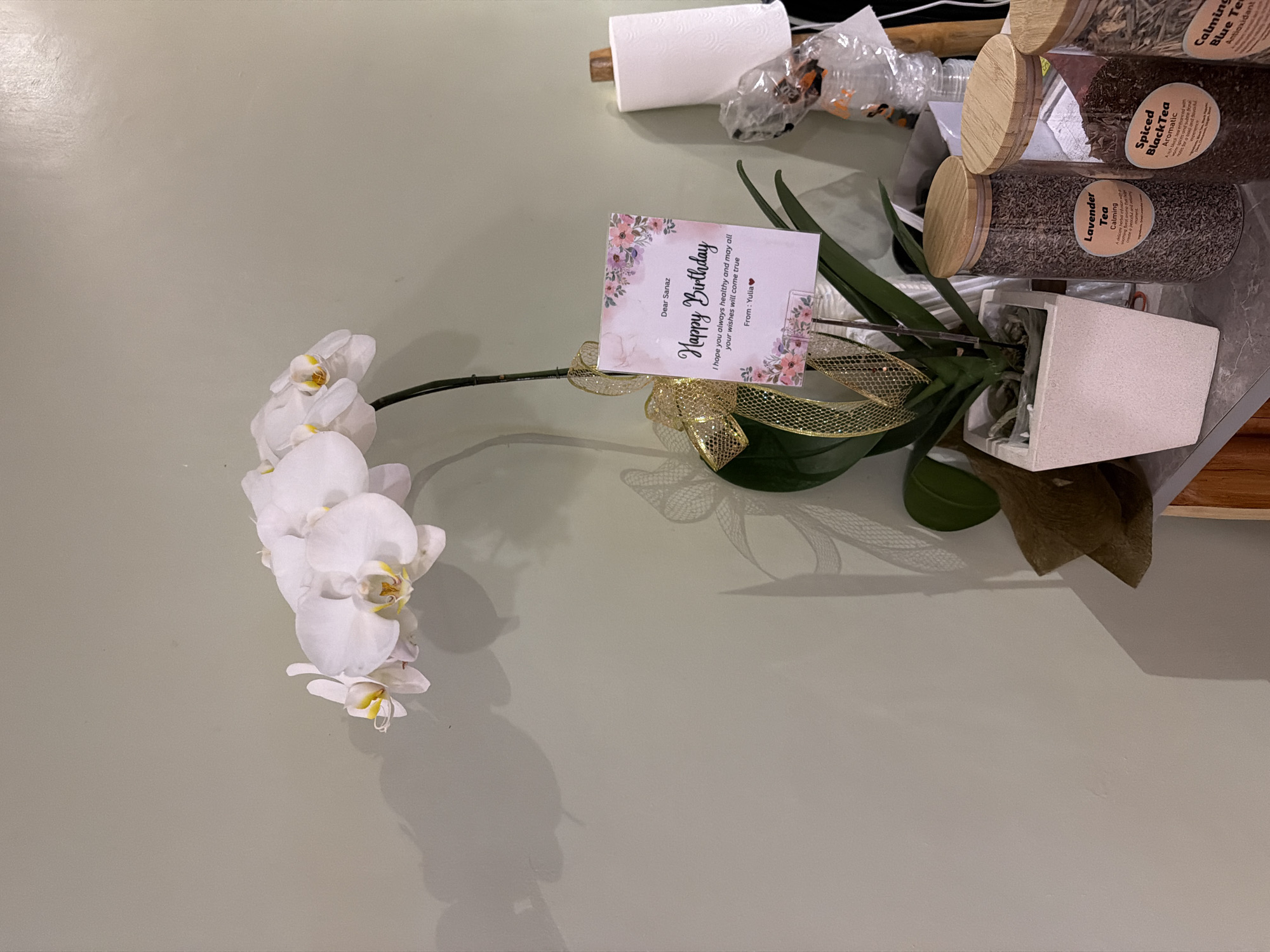 The image depicts a table with an orchid plant, a birthday card, and several canisters of tea. The orchid is white and in full bloom, extending across the left side of the table. A birthday card with the message "Happy Birthday" is attached to the plant with a gold ribbon. The card is addressed to Sanaz and signed by Yulia. To the right of the plant are several canisters of tea labeled as "Lavender Tea", "Spiced Black Tea", and "Calming Blue Tea". Other items such as a roll of paper towels and various plastic bags are visible in the background. The surface of the table is light green and appears to be in a domestic setting. The location is not evident in the image, therefore the association with North Kuta, Indonesia is unsupported by the provided context.