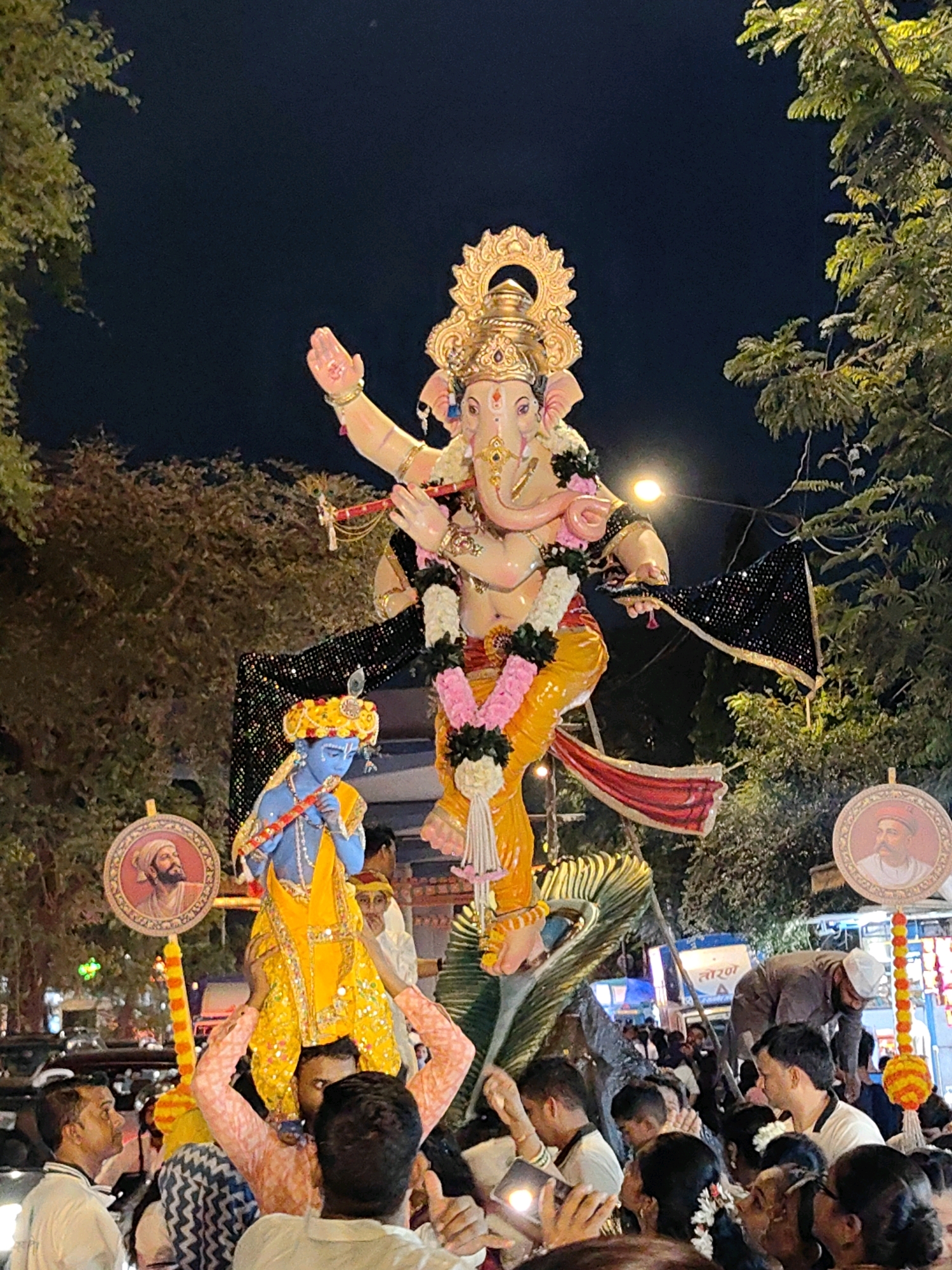 The image depicts a nighttime religious procession in Mumbai, India, likely celebrating Ganesh Chaturthi. A large, brightly colored idol of the deity Ganesha, adorned with garlands, is the central focus. To the left and slightly lower is a smaller idol of Krishna, with blue skin and yellow attire, holding a flute. Both idols are mounted on a platform, presumably carried by unseen individuals. The scene is crowded with people, many of whom are looking towards the idols. Some individuals are holding up phones to take pictures, indicating a public event. The procession appears to be taking place on a street, with trees lining the background. Decorative elements, including circular portraits of historical figures and colorful garlands, are visible. The overall atmosphere suggests a festive and spiritual gathering.