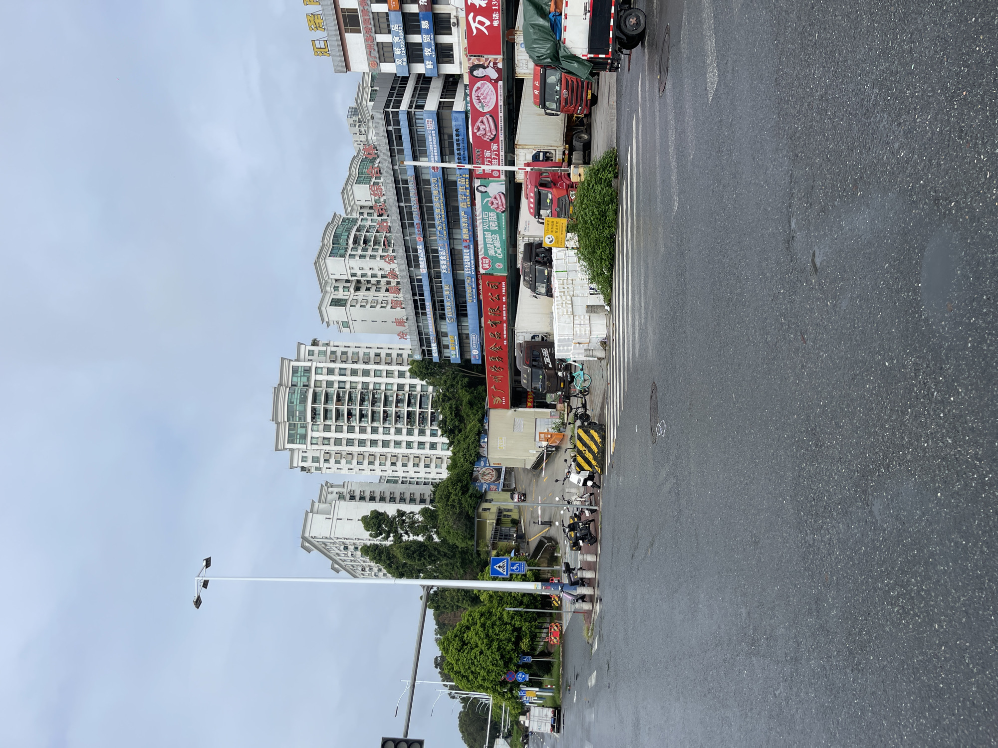 The image shows an outdoor urban scene in Panyu District, China, likely at an intersection or near a commercial area, with wet asphalt roads suggesting recent rain. In the foreground, a pedestrian crossing is visible, along with a yellow and black striped barrier. Several motorcycles and scooters are parked to the right of the barrier. Further back, multiple large trucks, including refrigerated trucks, are parked. Some trucks are white, and others are red. White boxes are stacked near some of the trucks.
Behind the trucks, a multi-story building complex is prominent. The lower levels of the building feature blue and white signage, some with Chinese characters. One sign, in red characters, appears to be advertising food products, possibly sausages, with images of sausages and people. Higher floors of the buildings are residential or office spaces, with numerous windows and balconies. Trees are visible between the buildings and at the edge of the road. A traffic light with black casings is visible on a pole on the left side of the image, along with a tall, white street lamp. The sky is overcast and grey. Several individuals are visible on the sidewalk to the left, appearing to be standing or walking. A blue and white traffic sign with a disabled person symbol is mounted on a pole near the parked motorcycles.