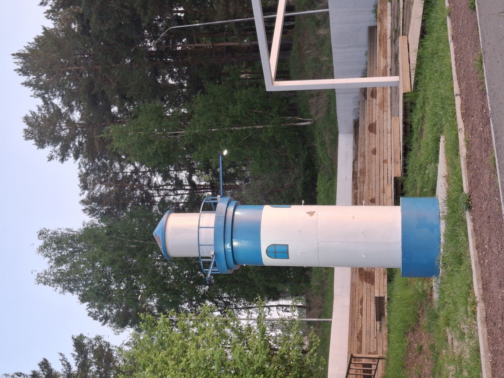 The image depicts a model lighthouse, colored white and blue, in a park setting. The lighthouse is positioned vertically in the image, appearing to stand on a green grass verge bordered by a walkway on one side and a wooden boardwalk on the other. Trees are visible in the background. A white, rectangular architectural structure is situated behind the lighthouse. The image is taken outdoors. Based on the prompt, the location is Chaikovsky, Russia.
