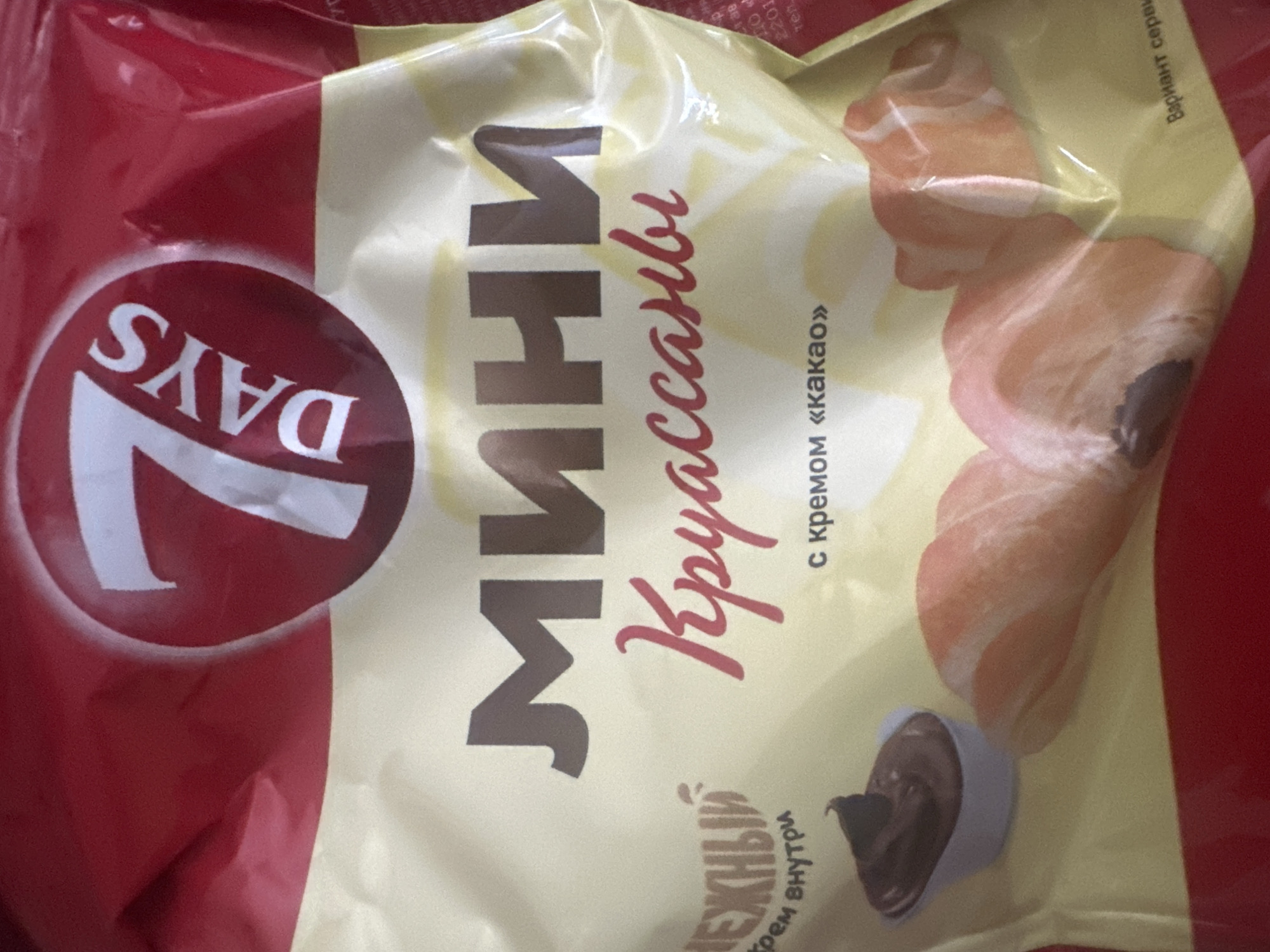 This detailed image depicts the front of a crinkled, flexible food package for mini croissants, located in the city of Unknown, Unknown. The packaging is predominantly red at the top and bottom sections, with a broad cream-colored band across the middle. A prominent circular red logo featuring a white number "7" and the word "DAYS" is situated in the upper right. Below this, the word "МИНИ" (MINI) is displayed in large, brown Cyrillic letters, followed by "Круассаны" (Croissants) in flowing red Cyrillic script. To the left, vertically aligned, the phrase "нежный крем внутри" (tender cream inside) is written in small brown Cyrillic text. An illustration of a small white bowl filled with dark brown chocolate cream is positioned in the lower-left portion of the cream band. To its right, several depictions of mini croissants are visible; one is partially cut open, revealing a dark filling, and another intact croissant is nearby. Below the main product name, in smaller brown Cyrillic text, is "с кремом «какао»" (with cocoa cream). A faint, patterned background is visible within the cream-colored section. The bottom right edge of the package shows partial Cyrillic text reading "Вариант сервировки" (Serving suggestion). No people, actions, or interactions are visible in the scene.
