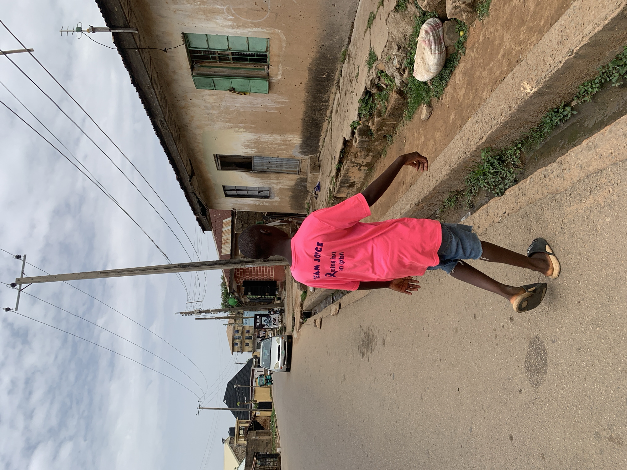 The image captures a street scene in Jos, Nigeria. A young person wearing a bright pink t-shirt with "Team Joyce" printed on the back walks along the side of a paved road. The t-shirt also has "Roning Init in opcion" written on it in pink and black, in all capital letters, with a pink ribbon above it. The individual is also wearing denim shorts and sandals, is walking towards the street's side of the scene. Adjacent to the road is a concrete gutter and a concrete sidewalk. On the other side of the sidewalk is a building with faded, peeling beige paint and green window shutters. A bag, white with dark stripes, is visible resting on the sidewalk. In the background, the street stretches away, showing other buildings, parked cars, and utility poles. The sky is overcast.
