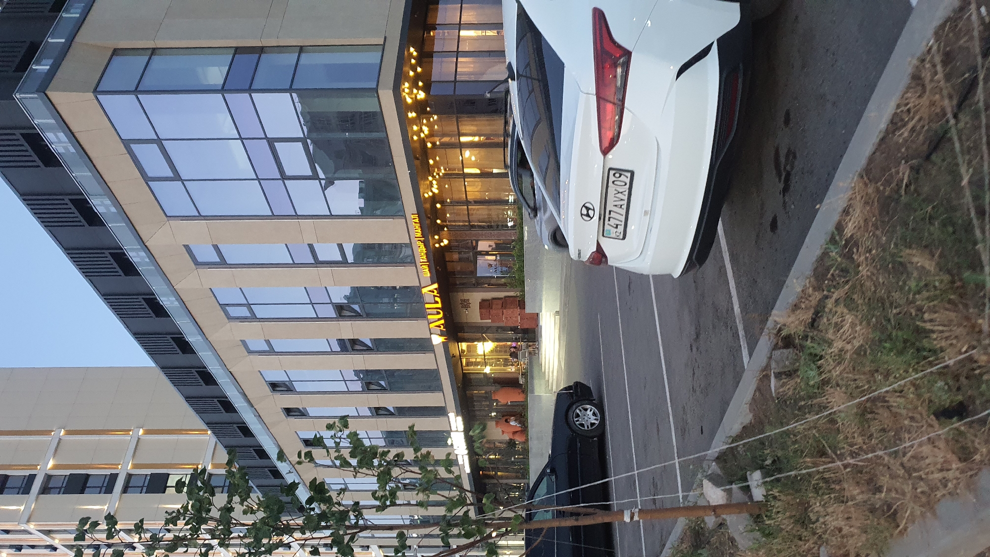 A white Hyundai sedan is parked in a parking lot in front of a modern building. The building has large glass windows and decorative lights along the facade. A black sedan is also visible in the parking lot. The area appears to be an urban environment, possibly in Qarağandı city administration, Kazakhstan, given the license plate on the Hyundai. The sky is clear and it appears to be daytime.