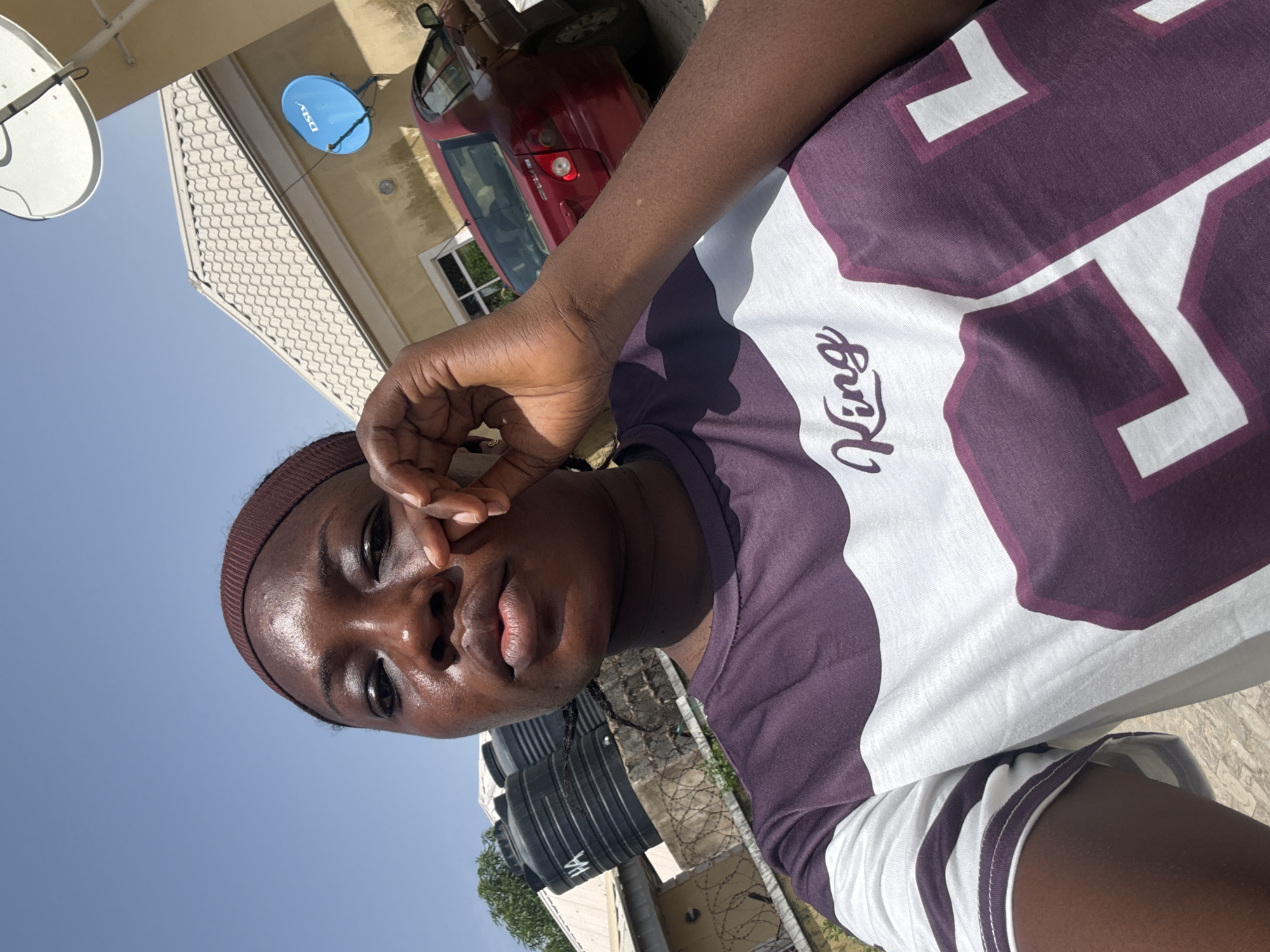 The image shows a person with dark skin, wearing a brown headband, looking at the camera with a hand near their face. The person is wearing a shirt with a design of large purple numbers on a white background and the word "King" written in script. In the background, there is a building with a satellite dish, a red car, black water tanks, and barbed wire. The location appears to be outdoors, in the city of Abuja, Nigeria, under a bright, sunny sky.