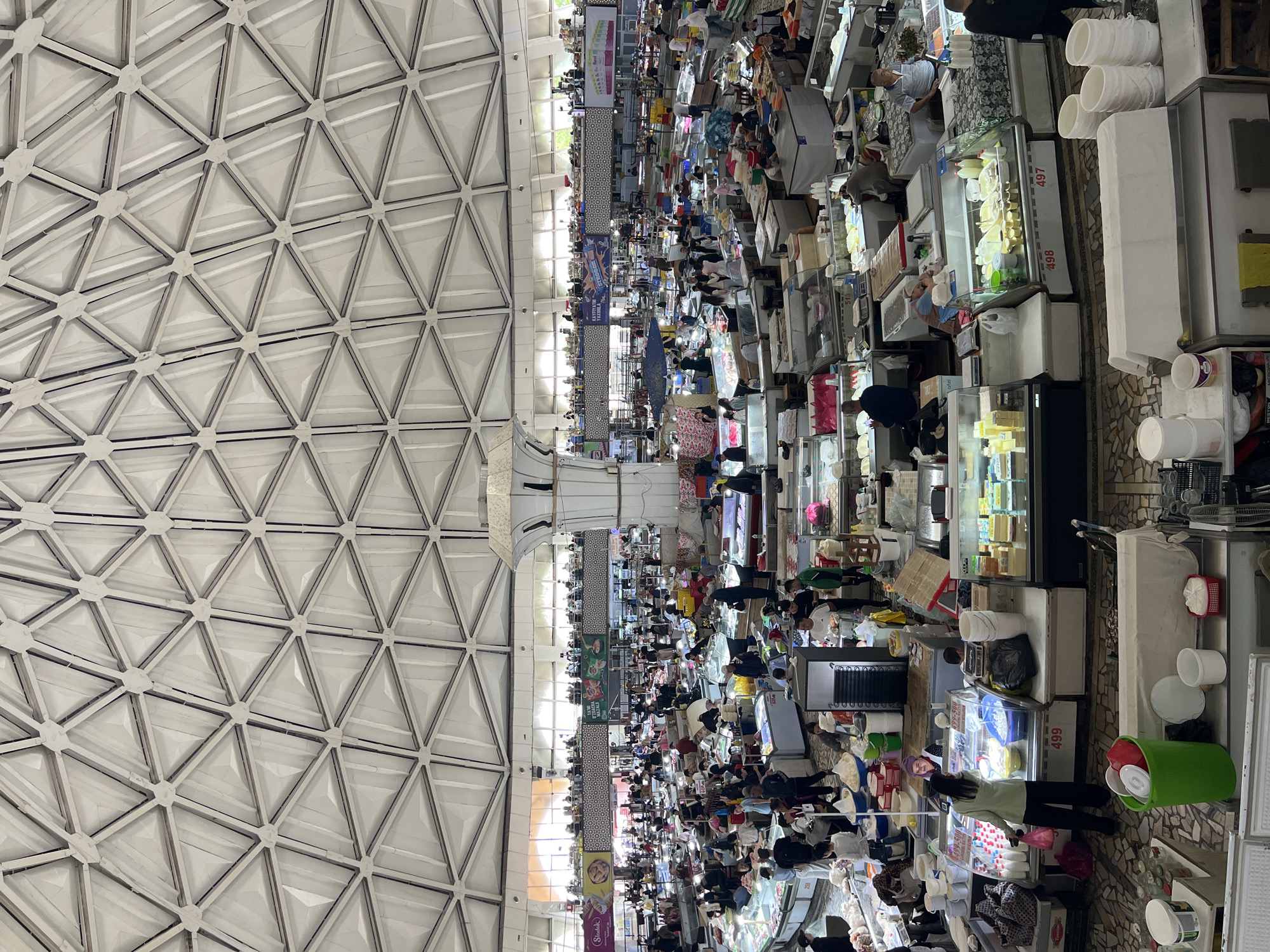 The image shows the Chorsu Bazaar in Tashkent, Uzbekistan, viewed from above. The bazaar is crowded with people milling between numerous stalls. Stalls are stocked with goods, including food items in refrigerated display cases and various other products. The ceiling is a white, triangulated pattern.