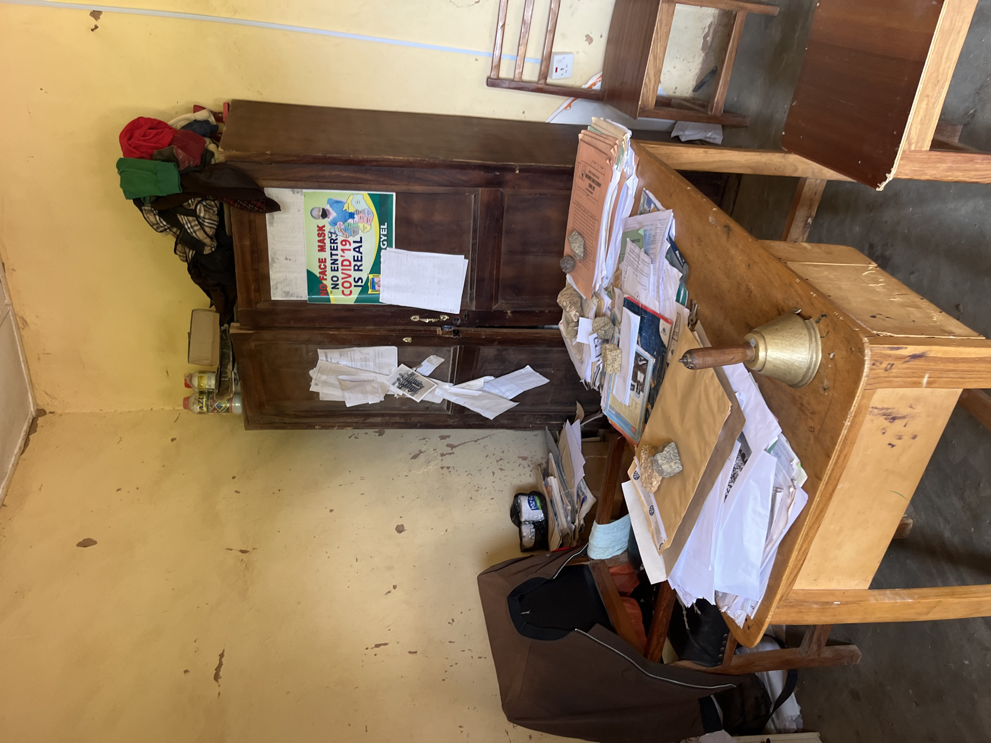 The image depicts a cluttered office space, likely a classroom or administrative office, in Bukuru, Nigeria. The room is furnished with a wooden desk piled high with papers, folders, and other documents, along with a brass bell. A large wooden cabinet stands against the wall, with clothes stacked on top. A poster about COVID-19 prevention is displayed on the cabinet door. A chair draped with a brown jacket is positioned near the desk, and another wooden chair is visible to the right of the image. The walls are yellow and show signs of wear and tear. The floor appears to be concrete. The overall impression is one of a functional but somewhat disorganized workspace.