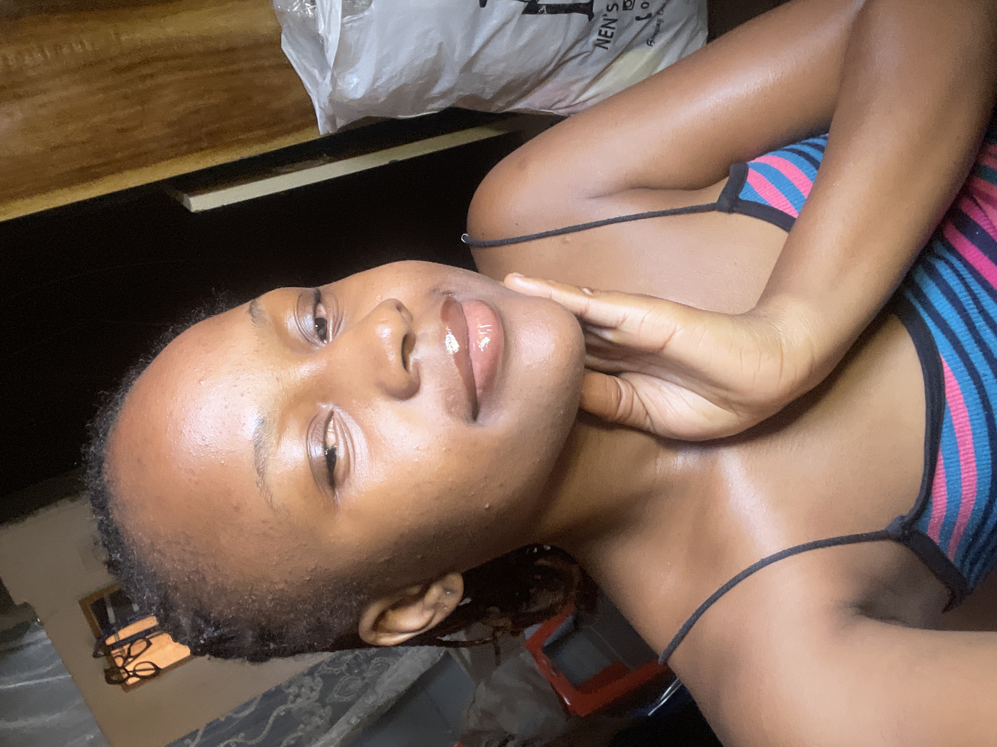 The image is a close-up selfie of a young Black woman with braided hair, wearing a multi-colored striped tank top. She is holding her right hand to her chin and looking at the camera with a slight smile. Her lips are glossy. The background is dimly lit and appears to be an indoor setting. To the left, there is a patterned curtain and a red chair. To the right, a large white bag with black lettering is visible. The lighting suggests it might be natural light coming from a window. The overall ambiance is casual and personal. The location is specified as the city of Jos, Nigeria.