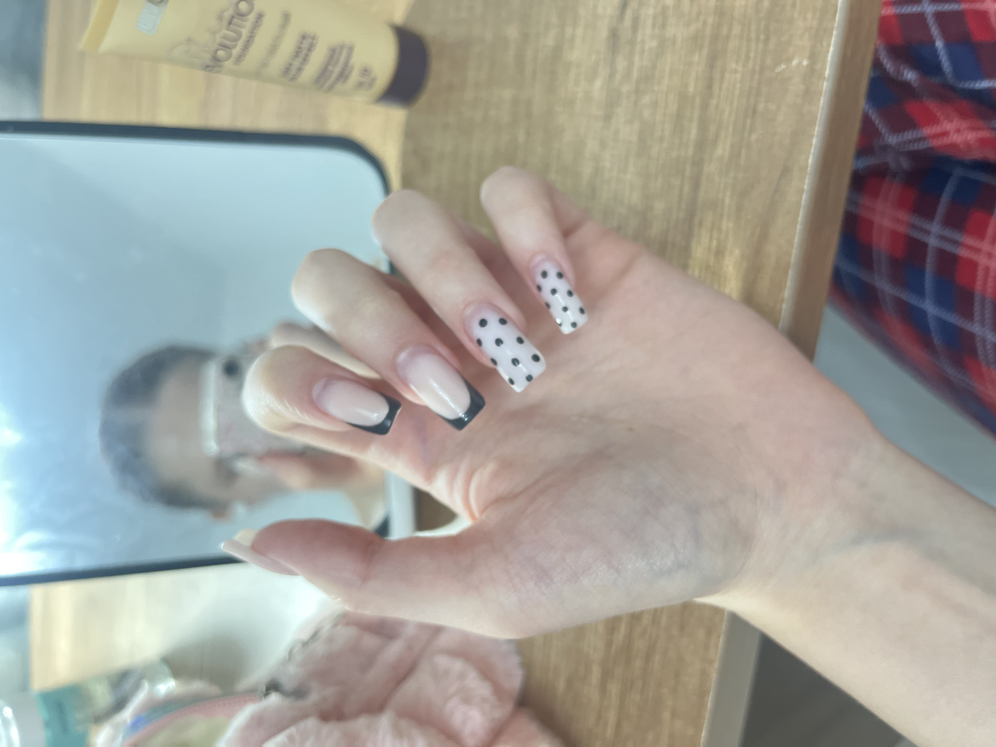 An eye-level close-up shot focuses on a person's left hand, showcasing an elaborate manicure, set against a light-toned wooden surface. The hand is angled with the palm facing downwards, displaying long, square-shaped artificial nails.
The nails feature varied designs: the thumb and index finger display a classic French manicure with a pale pink base and white tips. The middle finger's nail has a pale pink base with a distinct black French tip. The ring finger and pinky finger nails are adorned with a playful pattern of black polka dots over a pale pink base.
In the upper left quadrant of the image, partially obscured by the hand, a rectangular mirror reflects a blurred image of a person's head and shoulder, likely the individual taking the photo, along with indistinct elements of the surrounding room. On the wooden surface next to the mirror, a light-colored cosmetic tube, possibly foundation, is visible, with parts of the words "OLUTION" and "FOUNDATION" discernable on its label. To the right of the hand, a portion of red and blue plaid fabric is visible, suggesting clothing or a blanket. The scene appears to be an indoor setting, likely at a table or vanity, intended to highlight the intricate nail art. No specific time of day or emotions are apparent. The location context for this image is Perm, Russia.
