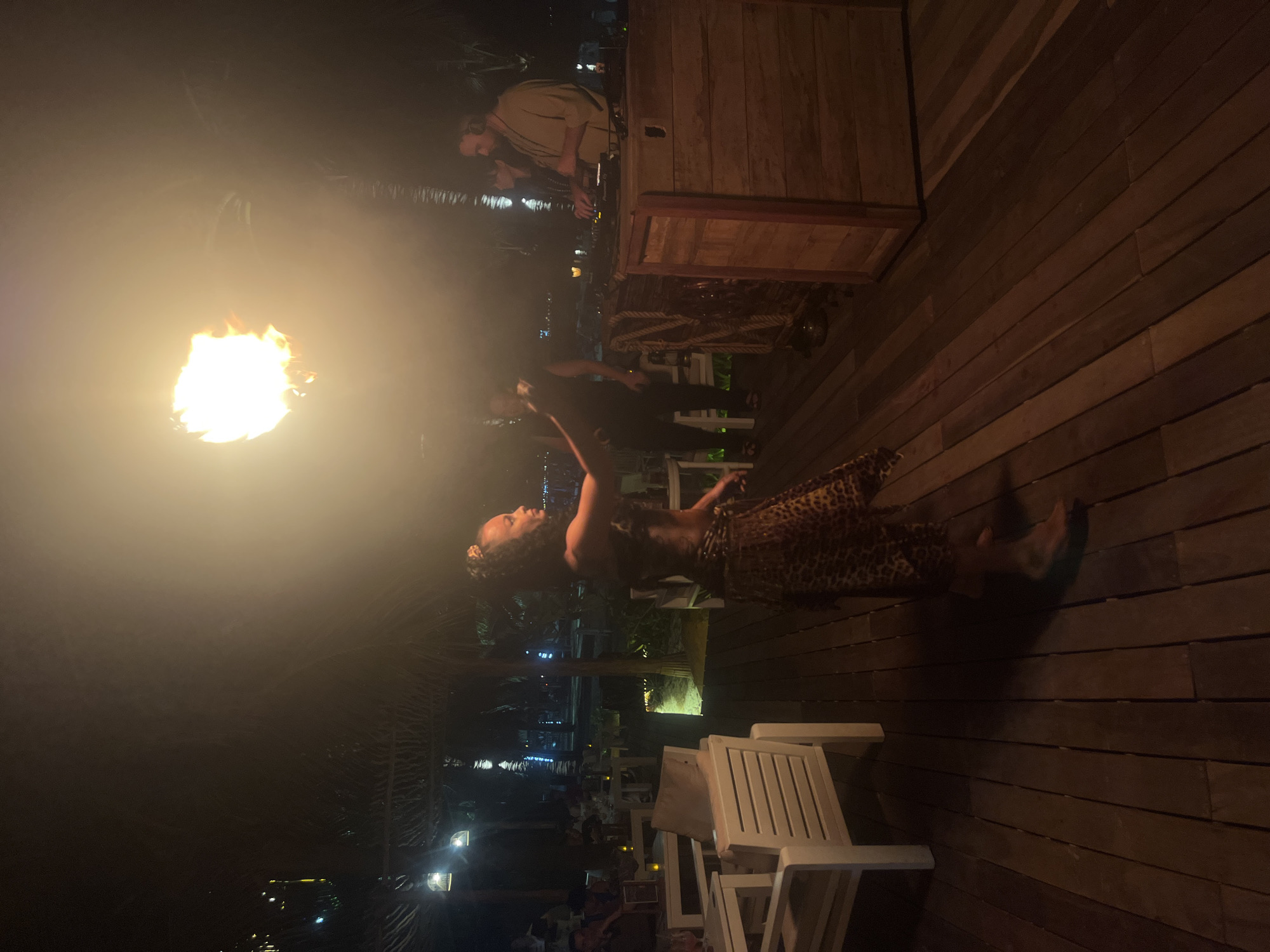 An outdoor nighttime scene at what appears to be a beachside resort or restaurant in Đà Nẵng, Vietnam. A woman in a leopard-print outfit is performing with a fire prop above her head. She is barefoot and her body is turned to her right. In the background, a DJ is seen operating equipment on a wooden console. Several white tables and chairs are visible, some with patrons seated. Palm trees and thatched roofs suggest a tropical setting. The ground is a wooden deck. The overall atmosphere is festive, with ambient lighting and the glow of the fire.