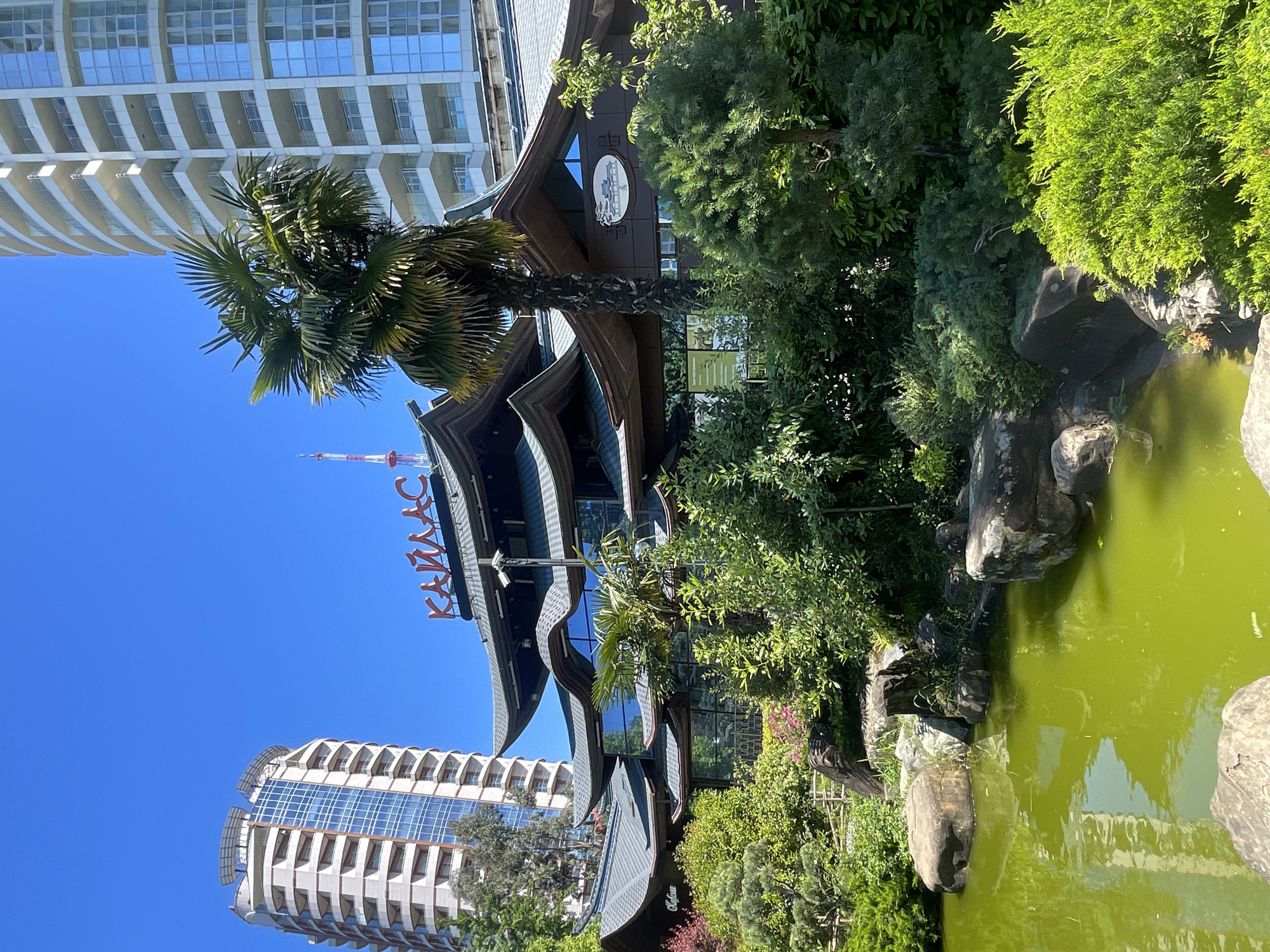 The image captures a vibrant outdoor scene under a clear blue sky, featuring a juxtaposition of modern architecture and a traditional East Asian-inspired building, surrounded by lush greenery and a tranquil pond.
In the mid-ground, a distinctive building stands out with multiple tiers of dark, curved roofs reminiscent of a pagoda, indicating an East Asian architectural style. This building has large windows and a prominent sign. Surrounding it and extending into the foreground on the right is a meticulously landscaped garden, dense with various green shrubs and trees, including several tall palm trees. Large, irregular grey boulders and rocks are artfully placed within the foliage and along the edge of a pond that occupies the right side of the foreground. The pond's water is a striking greenish hue, reflecting the clear blue sky and some surrounding elements.
In the background and to the left, two modern, tall high-rise buildings are visible. One prominent building has a light-colored, multi-story facade with numerous windows and balconies, some featuring elegant curved designs. Another, partially visible behind it, appears to be a typical office or residential building with a grid of reflective windows. In the far distance, a slender red and white antenna or mast pierces the sky. The bright, cloudless blue sky and strong illumination suggest it is a sunny day.
On the facade of the East Asian-style building, large red Cyrillic letters are clearly visible, spelling out "КАНАДС", likely part of a name for the establishment. No human activity is discernible, and the scene evokes a peaceful, resort-like atmosphere.