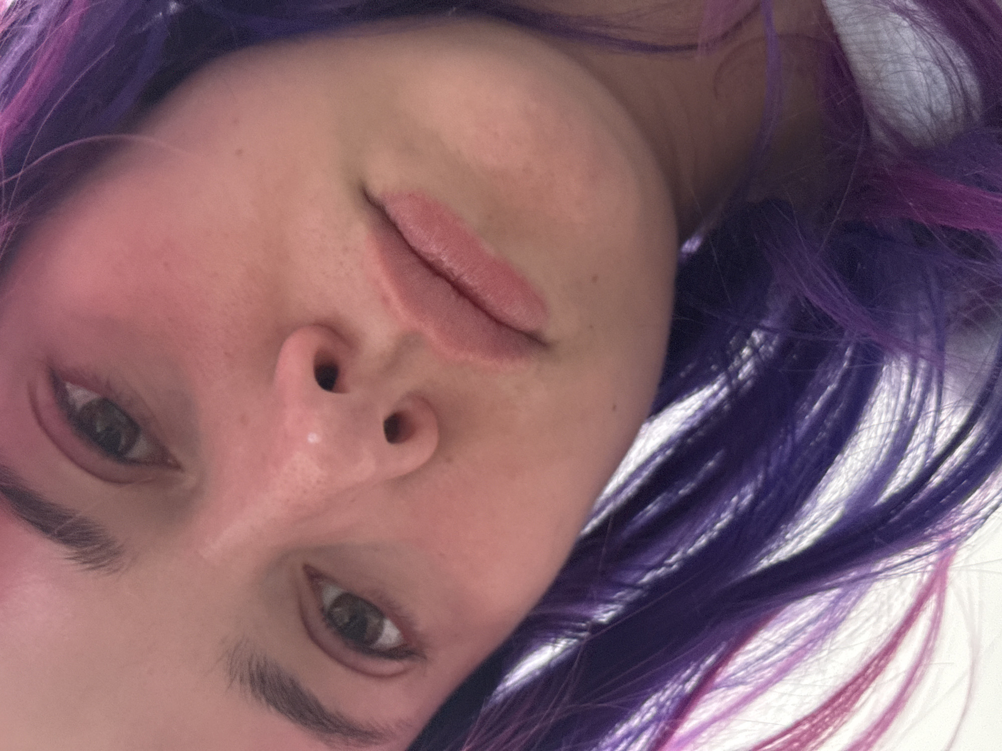 A close-up, upside-down view of a person's face with purple and pink hair. The skin is light-toned with visible pores and freckles. The eyes are brown, and the lips are pink. The background appears to be white. There is no information in the image to indicate this is in the city of Zagreb, Croatia.