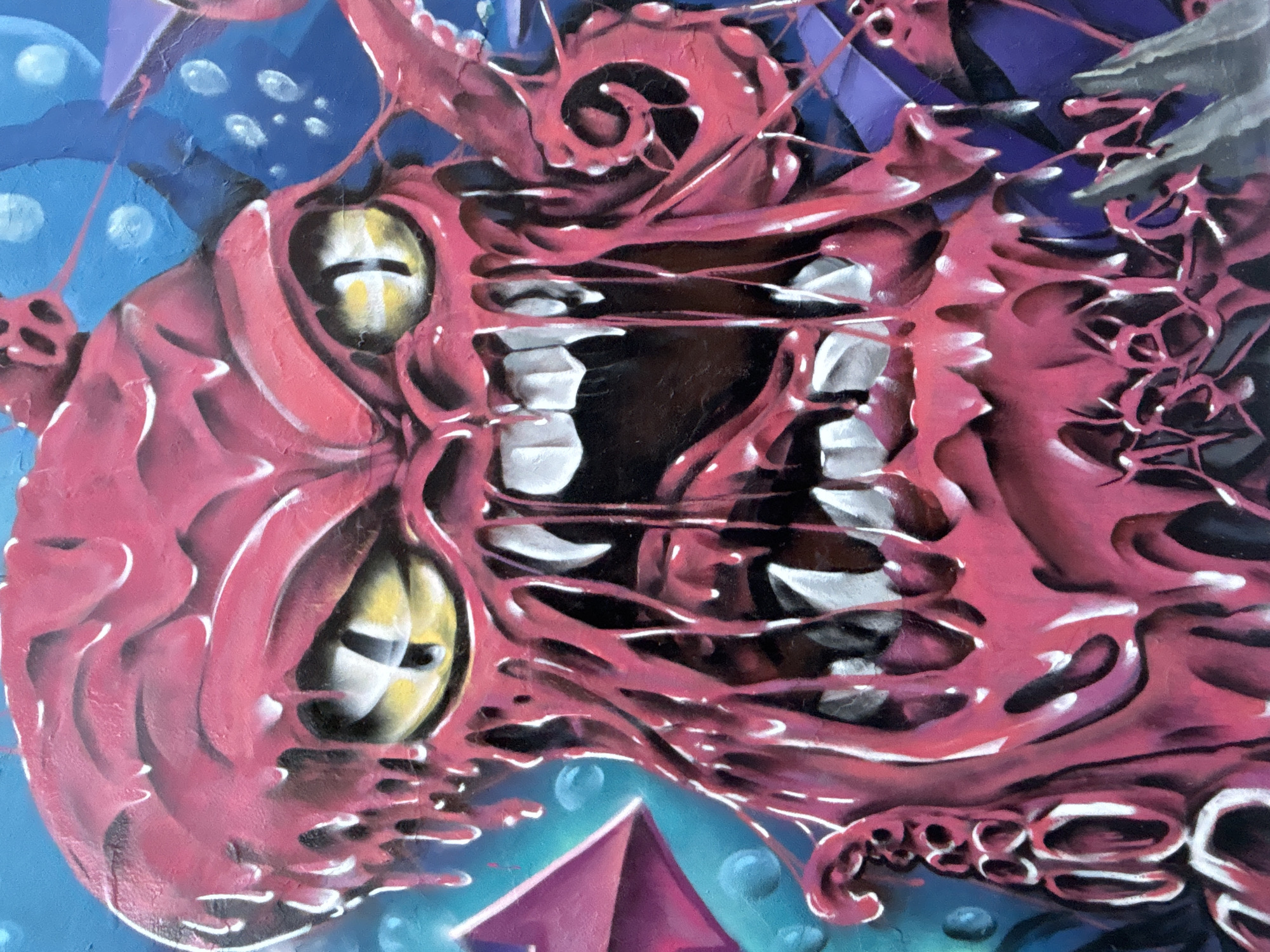 This vibrant image captures a close-up section of a stylized painting or mural, likely street art from Buenos Aires, Argentina. The central subject is a grotesque, monstrous creature rendered in vivid shades of pink and red, appearing fleshy and organic with prominent sinews and tendrils. It features two large, luminous yellowish-white eyes with dark, horizontal pupils, and a gaping maw filled with sharp, white teeth. The creature's expression suggests aggression, a primal roar, or a predatory snarl.
The background transitions from deep blues in the upper left to darker purples on the right, interspersed with scattered white, circular elements resembling bubbles or distant stars, hinting at an underwater or cosmic setting. In the lower portion, a striking, crystalline structure, possibly a pyramid or gem, appears in shades of purple and teal, also surrounded by smaller bubbles.
The artwork exhibits a distinct graffiti style, characterized by bold lines, exaggerated features, and a rich, saturated color palette. Highlights and shadows are strategically used to create a sense of depth and texture, particularly on the creature's textured skin and glistening teeth. There are no human subjects or visible text. The overall atmosphere is one of fantasy, horror, and surrealism, evoking a sense of awe and unease through its detailed and menacing depiction.