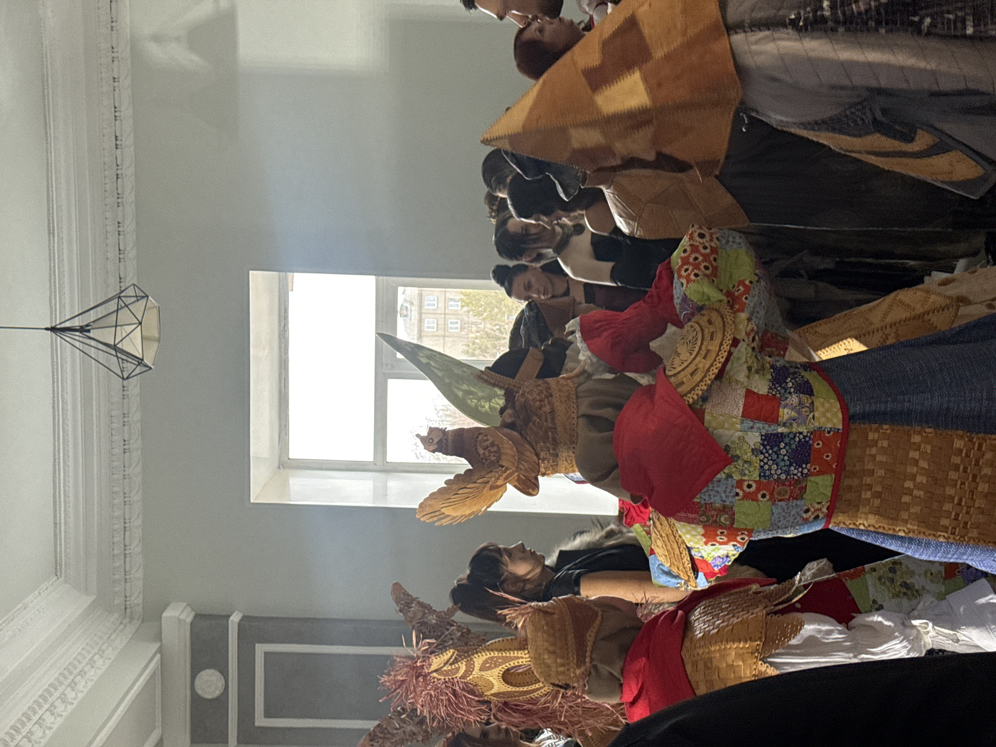 The image depicts a group of people indoors, possibly in a ceremonial setting. The location is suggested to be in the city of Unknown, Unknown. Some individuals are wearing traditional or artistic headwear, including one resembling a bird. The setting appears to be a room with a window and decorative elements on the walls and ceiling.