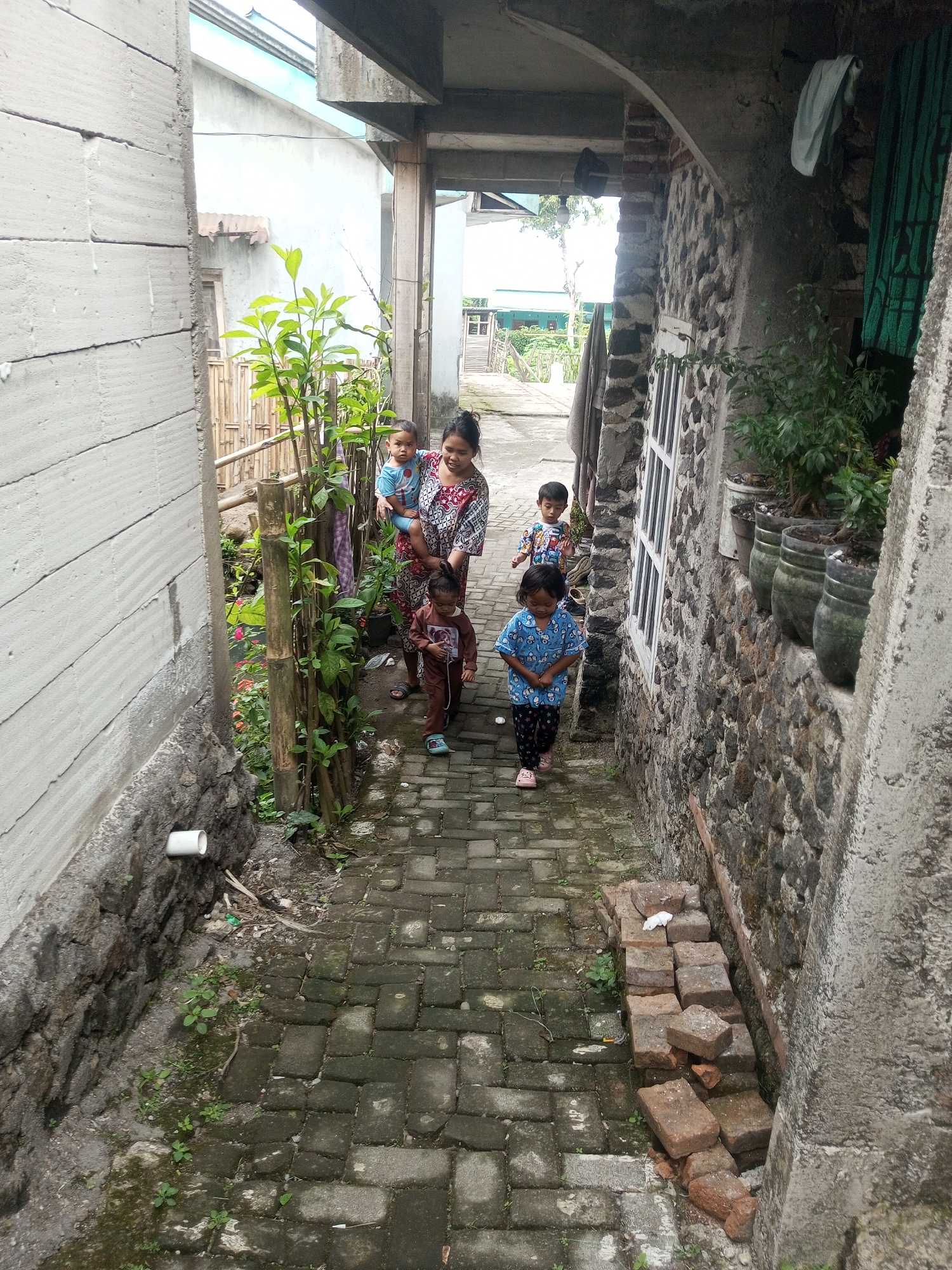 A mother is walking down a narrow, stone-lined alley in Andongsili, Indonesia, holding a baby and followed by three young children. The alley is paved with interlocking bricks, and a stone wall covered in moss runs along the right side. Several potted plants are arranged on a ledge above the wall. On the left, a lighter colored wall with horizontal grooves is visible, and further down, a bamboo fence encloses greenery. The lighting suggests it is daytime, with soft light filtering into the alley. The mother is wearing a patterned dress, and the children are in casual clothing. The scene depicts a family navigating a residential passage.