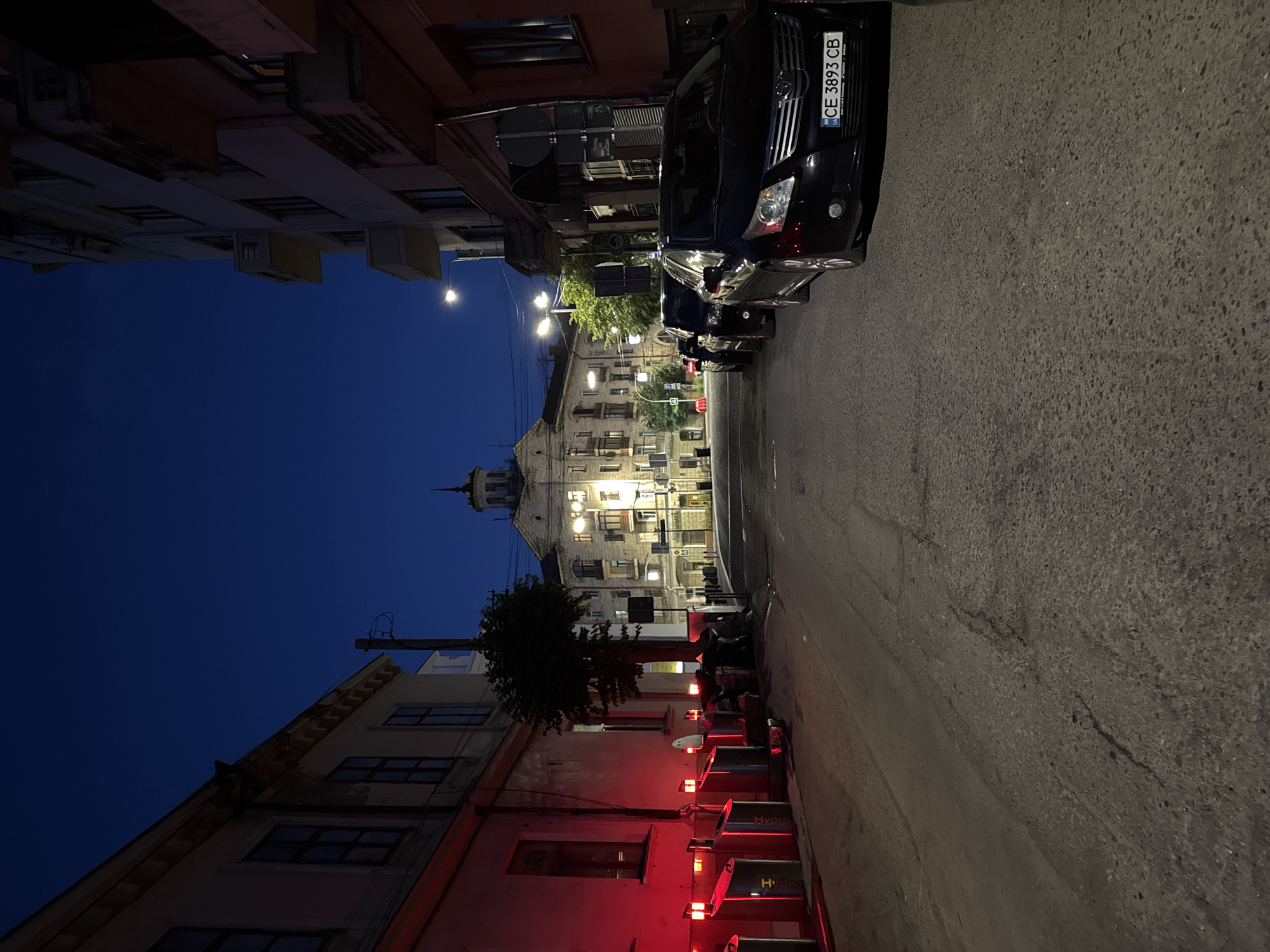 The image depicts a street scene at dusk in Chernivtsi, Ukraine. On the left side of the street, a building with several windows and red lighting illuminating the lower facade is visible. In front of this building, a row of modern, stainless-steel trash receptacles with red lights on top are arranged. A thin tree stands between the building and the street. Across the street, a line of parked black cars is visible, with the closest car featuring a license plate reading "CE 3893 CB". In the background, a large, ornate building with multiple stories and illuminated windows dominates the view, capped with a distinctive tower structure. Streetlights cast a glow on the road and sidewalk. The sky above is a deep blue, indicating twilight.
