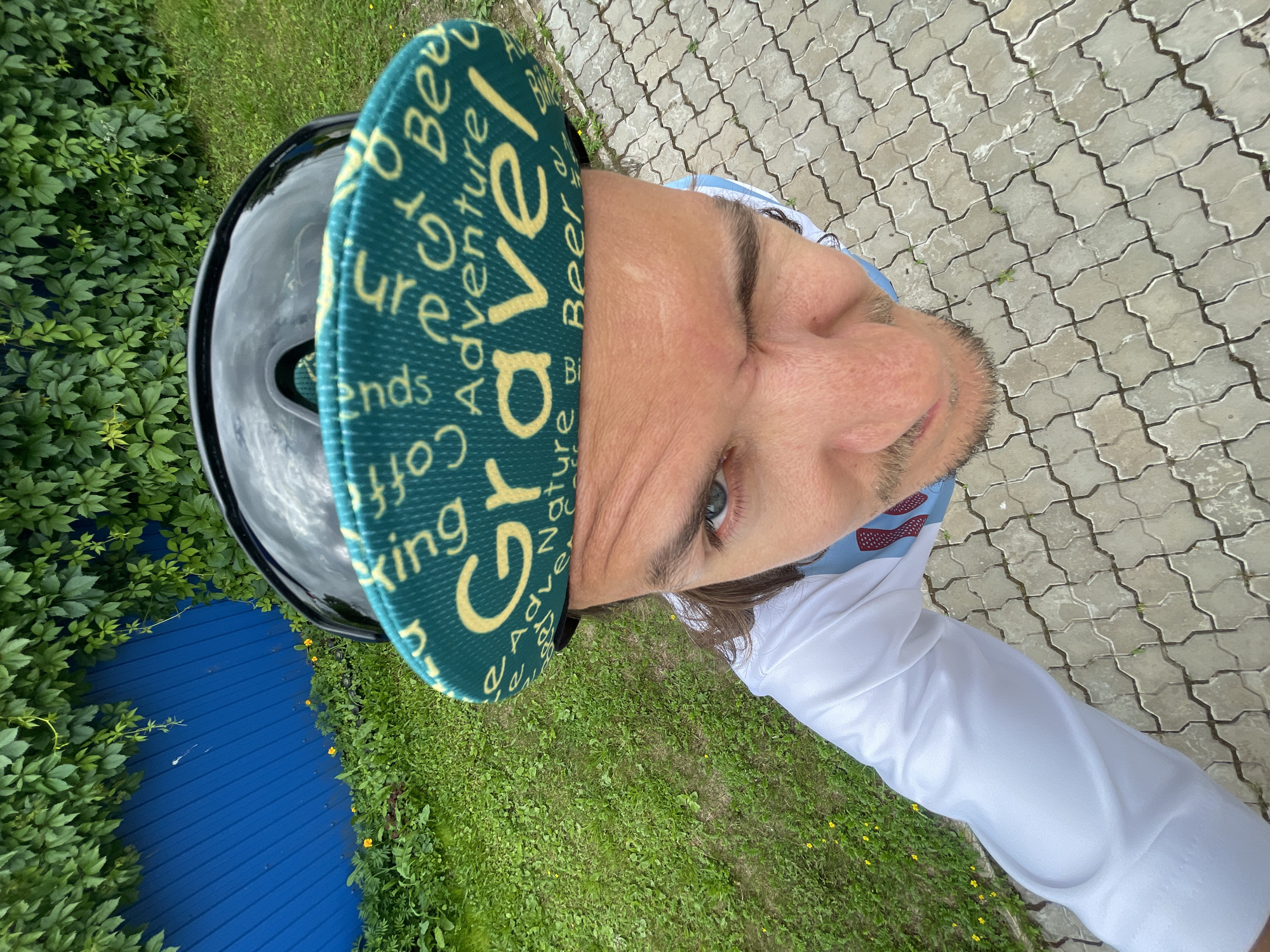 A man, wearing sunglasses and a cycling cap, is captured in a fisheye lens photograph taken from above. The cap displays the word "Gravel" in yellow lettering against a teal background, surrounded by other words like "Adventure," "Nature," and "Beer." He is also wearing a white shirt with a blue and red patterned collar. The background shows a vibrant blue wooden fence, overgrown with lush green ivy, and a grassy area with scattered yellow flowers. The ground is paved with interlocking grey concrete blocks. The man's expression is playful, as he winks at the camera.