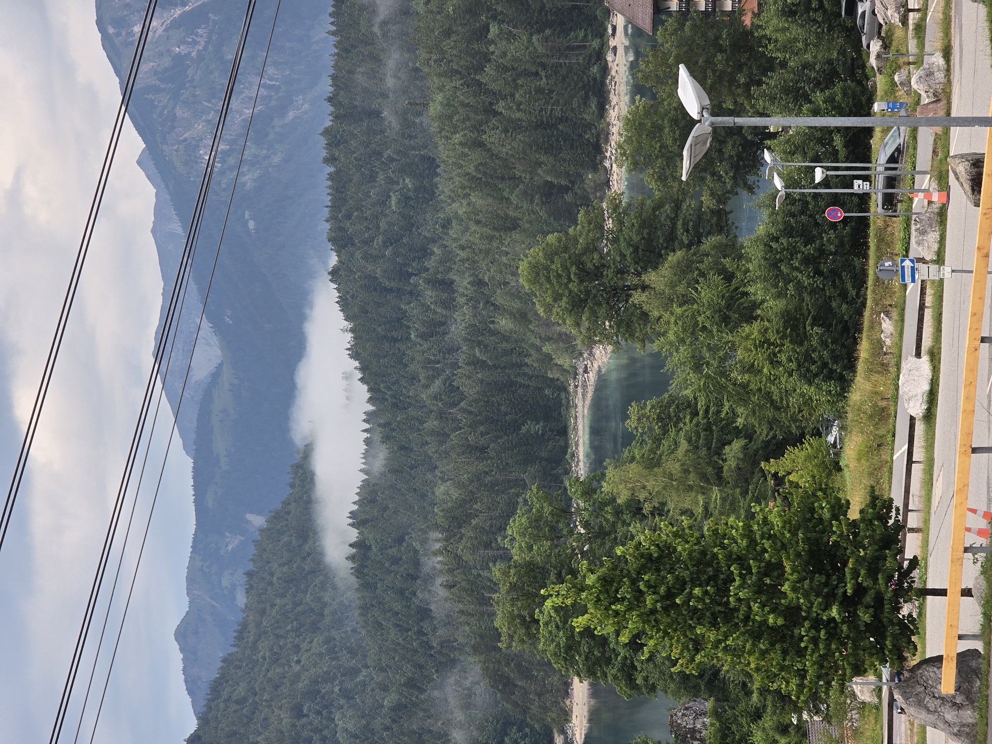 The image shows a scenic view of a lake surrounded by a dense forest, with mountains in the background. The sky is overcast, with some patches of fog or clouds obscuring the higher peaks of the mountains. In the foreground, there is a paved area with a road, a wooden fence, and several large rocks. Two modern streetlights stand on the right side of the frame, along with some traffic signs and a few parked cars. A building with a dark roof is visible on the far right. The overall scene suggests a natural landscape near a developed area, possibly a tourist destination. The location is identified as Grainau, Germany.