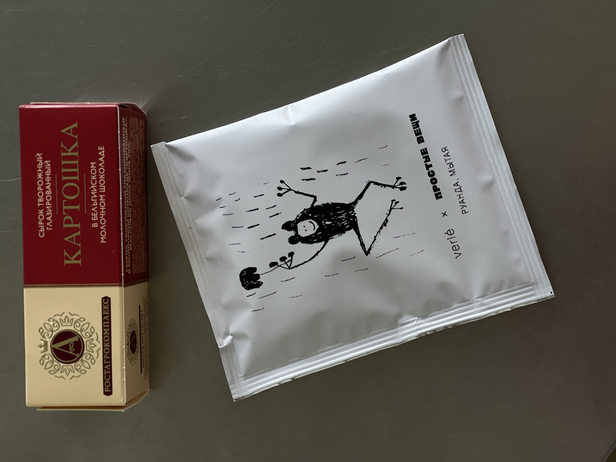 The image shows a close-up, overhead view of two food items placed on a gray, flat surface.
On the left is a small, dark red cardboard box with gold and white lettering. The text on the box is in Russian and appears to describe a dairy product, possibly a glazed curd snack, and mentions "Belgian milk chocolate." There's also a gold emblem with the letter "V" inside a decorative circle and the text "РОСТАГРОКОМПЛЕКС" below it.
To the right of the box is a white, foil-like packet with a black drawing of a frog-like creature seemingly swimming or moving through dashes. The packet also has text in Russian and French. The Russian text reads "ПРОСТЫЕ ВЕЩИ," which translates to "Simple Things." Below this, it says "РУАНДА, МЫТАЯ," indicating "Rwanda, Washed," likely referring to coffee. The French text says "verlé."
The lighting suggests it could be indoor lighting. There are no people or animals present, only the packaged food items. The overall impression is of a still-life product shot.