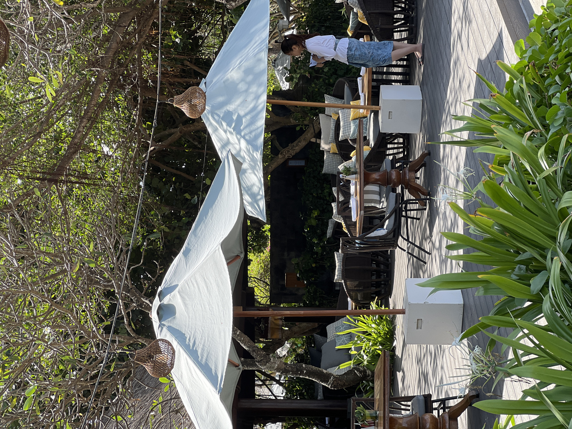 An outdoor seating area at a restaurant in Denpasar, Indonesia, is depicted in this medium-angle, eye-level shot. Two large, light blue umbrellas provide shade over wooden tables and rattan chairs. Lush green foliage frames the foreground, with a white flower visible amongst the leaves. In the background, more trees and a stone wall can be seen. A woman, wearing a white top and a blue patterned skirt, stands near one of the tables, looking at her phone. The ground is a wooden deck.