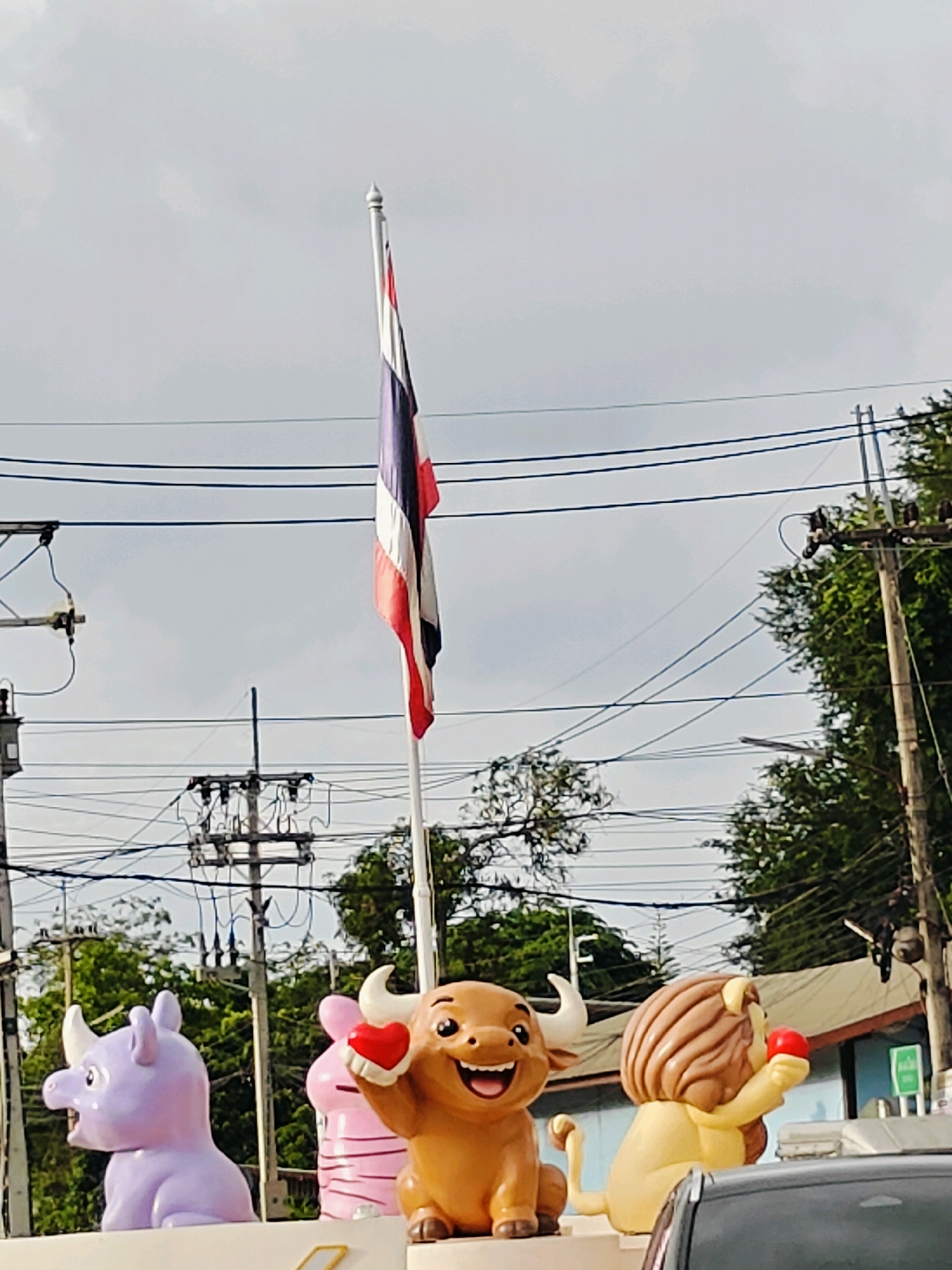 In the Tambon Mu Si, Thailand, an outdoor scene features several cartoon-like animal statues in the foreground. From left to right, a light purple rhinoceros statue with its mouth open and a horn is visible. Next to it is a pink pig statue holding a red heart. To the right of the pig, a light brown bull statue with white horns smiles broadly and holds a red heart. Further to the right, a yellow lion statue with a mane is depicted holding a red ball. Behind these statues, a flagpole with the Thai flag is prominently displayed against a cloudy sky. A complex network of electrical wires and utility poles stretches across the middle ground, with lush green trees and vegetation visible on the right side of the image. A building with a light-colored roof is partially visible behind the statues and trees. The overall impression is a bright, somewhat overcast day in a semi-urban or roadside setting.