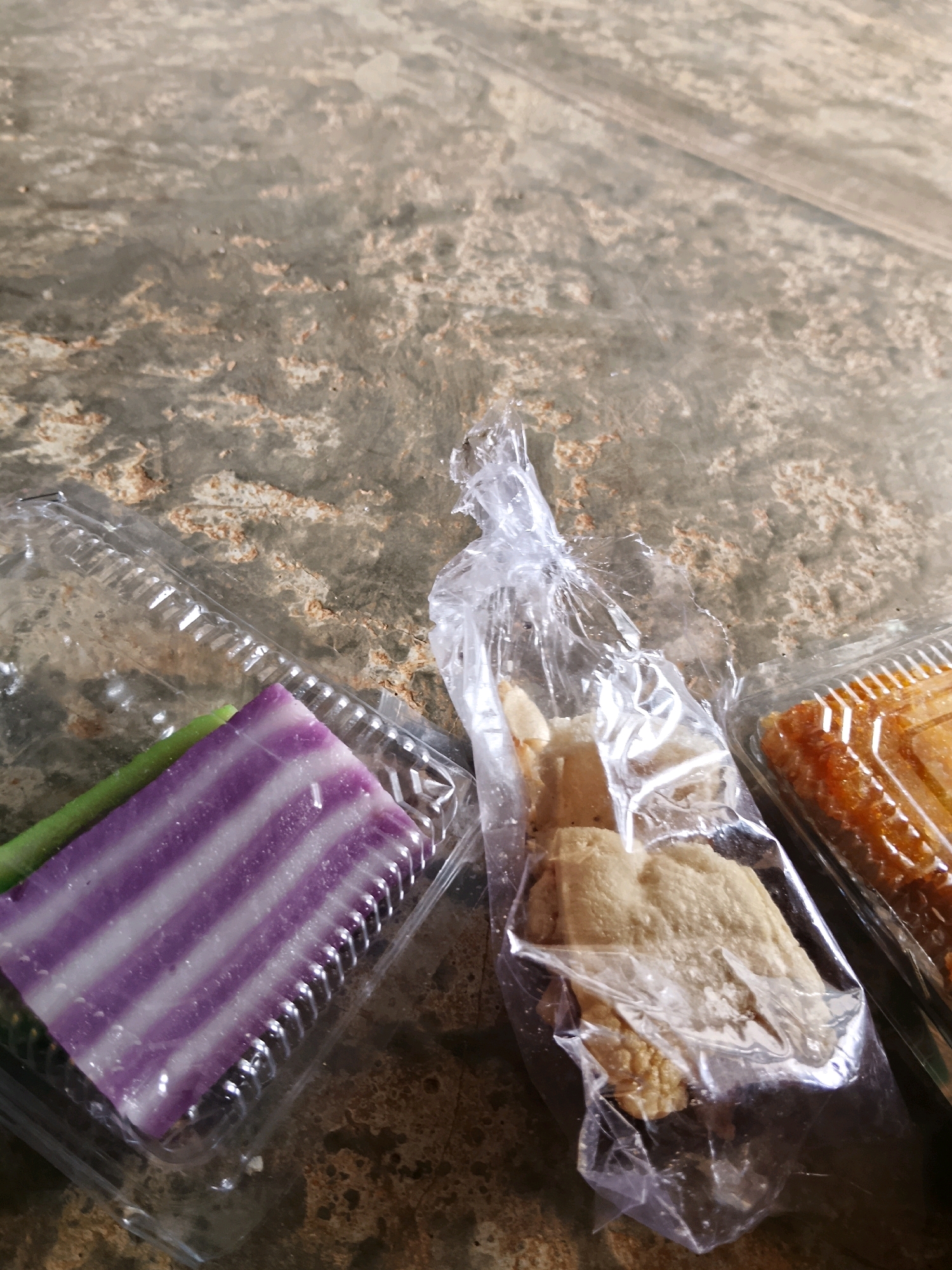 Three small food items in clear plastic packaging are arranged on a mottled, grey-brown surface. The item on the left is a rectangular piece of purple and white layered cake or dessert, with a green stick-like object visible next to it. The middle item consists of several irregular, light-brown pieces of food in a loosely tied plastic bag. The item on the right is a rectangular piece of what appears to be a golden-brown pastry or sweet in a clear plastic container. The surface beneath the food items has a rough, textured appearance with variations in color.