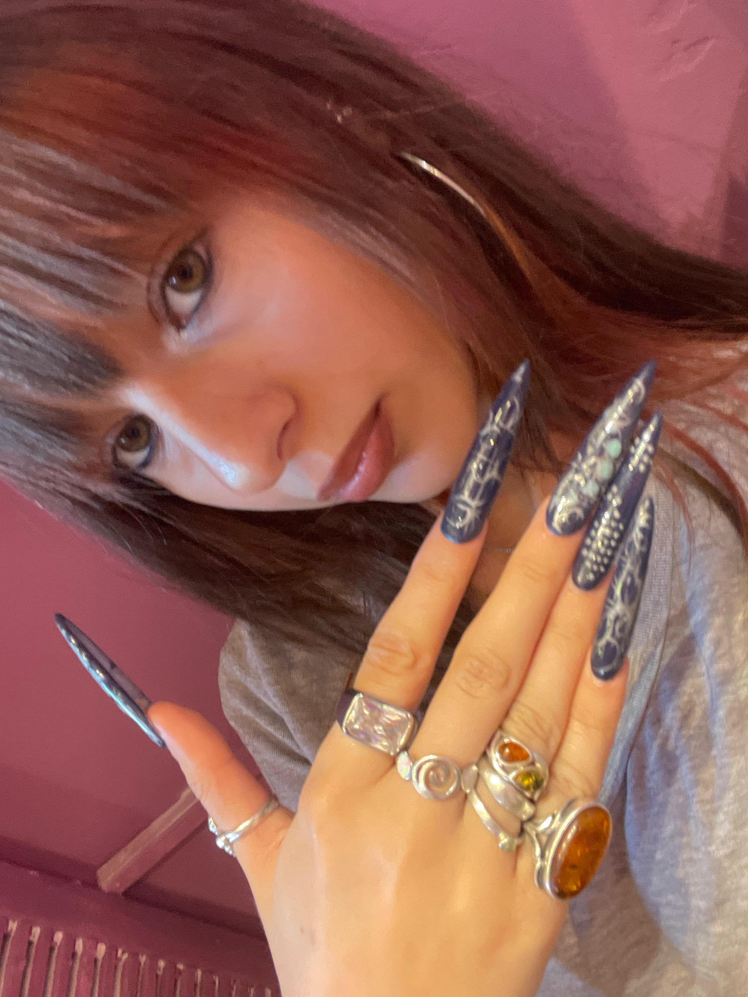 The image shows a close-up of a young woman's face and hand, with a pink background. She has long, dark hair with some reddish highlights, and bangs. Her right hand, adorned with several silver rings and long, pointed dark blue nails with intricate white designs, is raised towards her face. The background appears to be a wall or curtain with a textured surface, possibly a fabric or painted material. The lighting is soft, creating a slightly reflective sheen on her skin and nails. There are no discernible landmarks or objects that would indicate a specific location within Raanana, Israel.