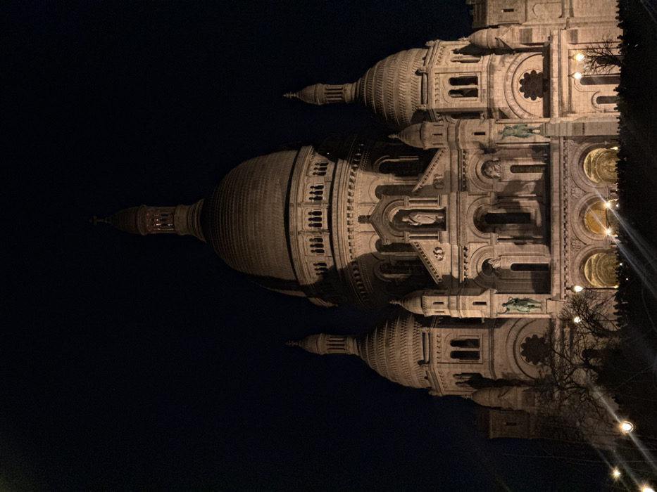 The image displays the Sacré-Cœur Basilica in Paris, illuminated at night. The sky is completely dark, suggesting late evening or night. The building is made of light-colored stone, which appears warm yellow and beige due to the artificial lighting.
The basilica features a prominent, large central dome with a tall spire on top, detailed with windows and architectural flourishes. To either side of the central dome, there are smaller, intricately designed domes, also topped with spires. The facade of the building is highly ornate, characterized by multiple arches, columns, and decorative carvings. Several statues are visible in niches along the facade, particularly noticeable on the upper levels. Circular and rectangular windows with detailed frames are interspersed across the structure. The lighting highlights the intricate textures and three-dimensional elements of the architecture, casting soft shadows that emphasize its depth.
On the far right side of the image, the dark silhouettes of bare tree branches are visible, contrasting against the illuminated building. Below these branches and extending into the lower right corner, a string of warm, glowing light bulbs is present, possibly part of a decorative lighting arrangement in a nearby park or pathway. The image is taken at eye level or slightly from below, emphasizing the grandeur of the basilica against the dark sky. The overall impression is one of serene beauty, with the historic landmark bathed in a golden glow.
(Note: The original image is oriented vertically, but the building itself is rotated 90 degrees clockwise, making it appear to be lying on its side. The description above treats the building as if it were correctly oriented vertically, with the domes at the top).