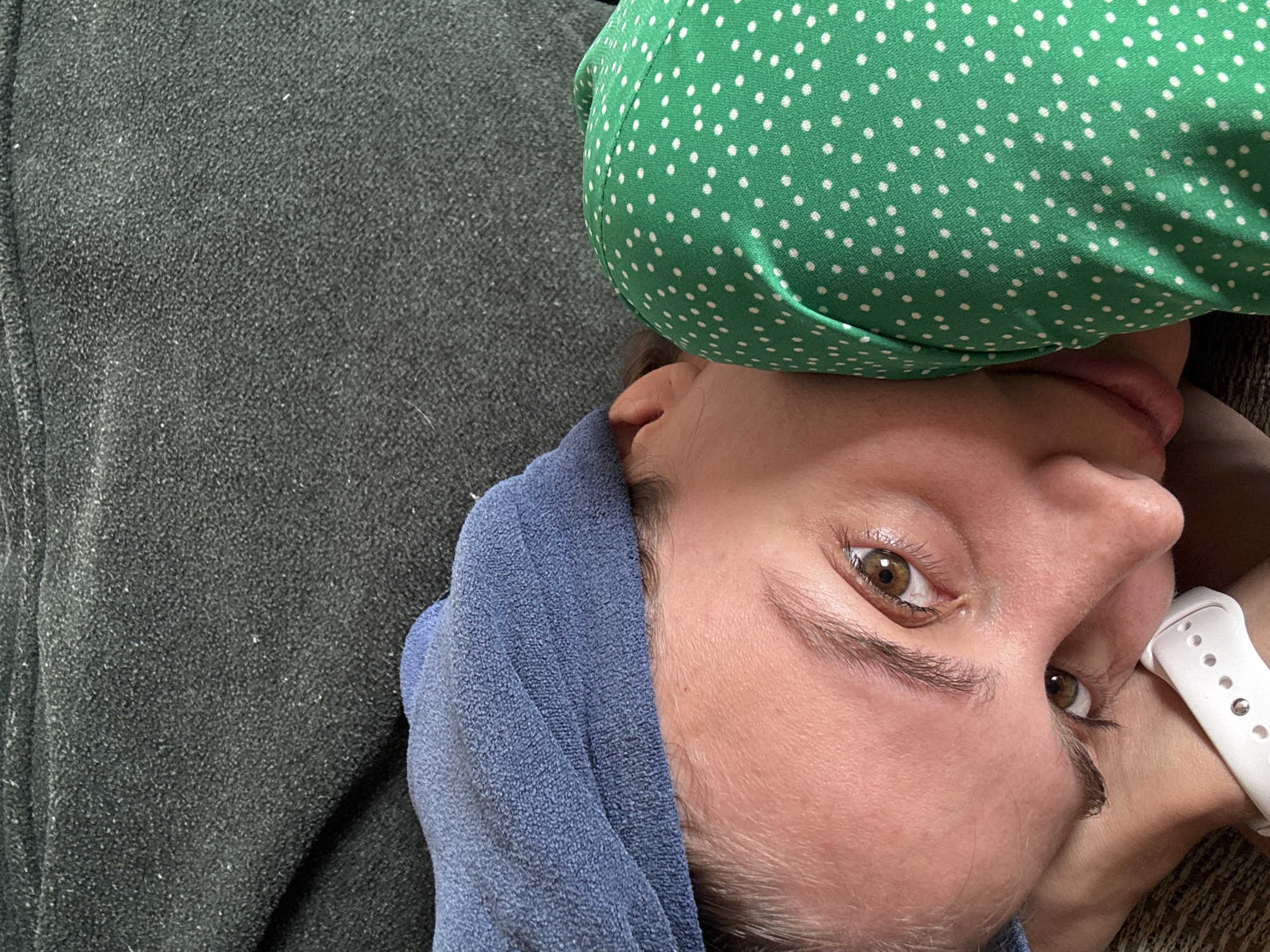 The image presents a close-up, high-angle, and tilted shot of a person, likely a woman, lying down. Her head is turned to the side, with her gaze directed towards the viewer.
The subject has fair skin, prominent brown eyes with visible eyelashes and eyebrows. Her lips are slightly parted. A white smartwatch with a perforated band is worn on her left wrist, positioned near her face.
She is wearing a vibrant green garment adorned with small white polka dots, which is visible on the upper right side of the frame and drapes over her body. A soft, blue textured fabric, resembling a towel or fleece material, is wrapped around her head or neck, partially covering her left side.
The majority of the background consists of a dark grey or black textured fabric, likely a blanket, clothing, or upholstery, which fills the left and lower portions of the image. This fabric has a fleecy appearance and shows minor lint or dust.
The setting appears to be indoors, suggesting a relaxed environment such as a couch or bed. The lighting is soft, highlighting the textures of the various fabrics and the details of the person's face. No discernible text is visible within the image.