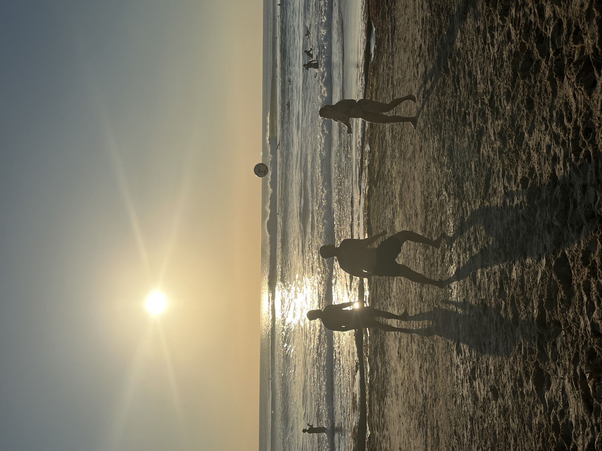 At sunset on Kuta Beach, Bali, Indonesia, three silhouetted figures are playing volleyball on the sandy shore. The sun, a bright orb low in the sky, casts long shadows and reflects brilliantly on the ocean waves. In the background, other beachgoers and surfers can be seen scattered across the water and sand, enjoying the warm evening light.