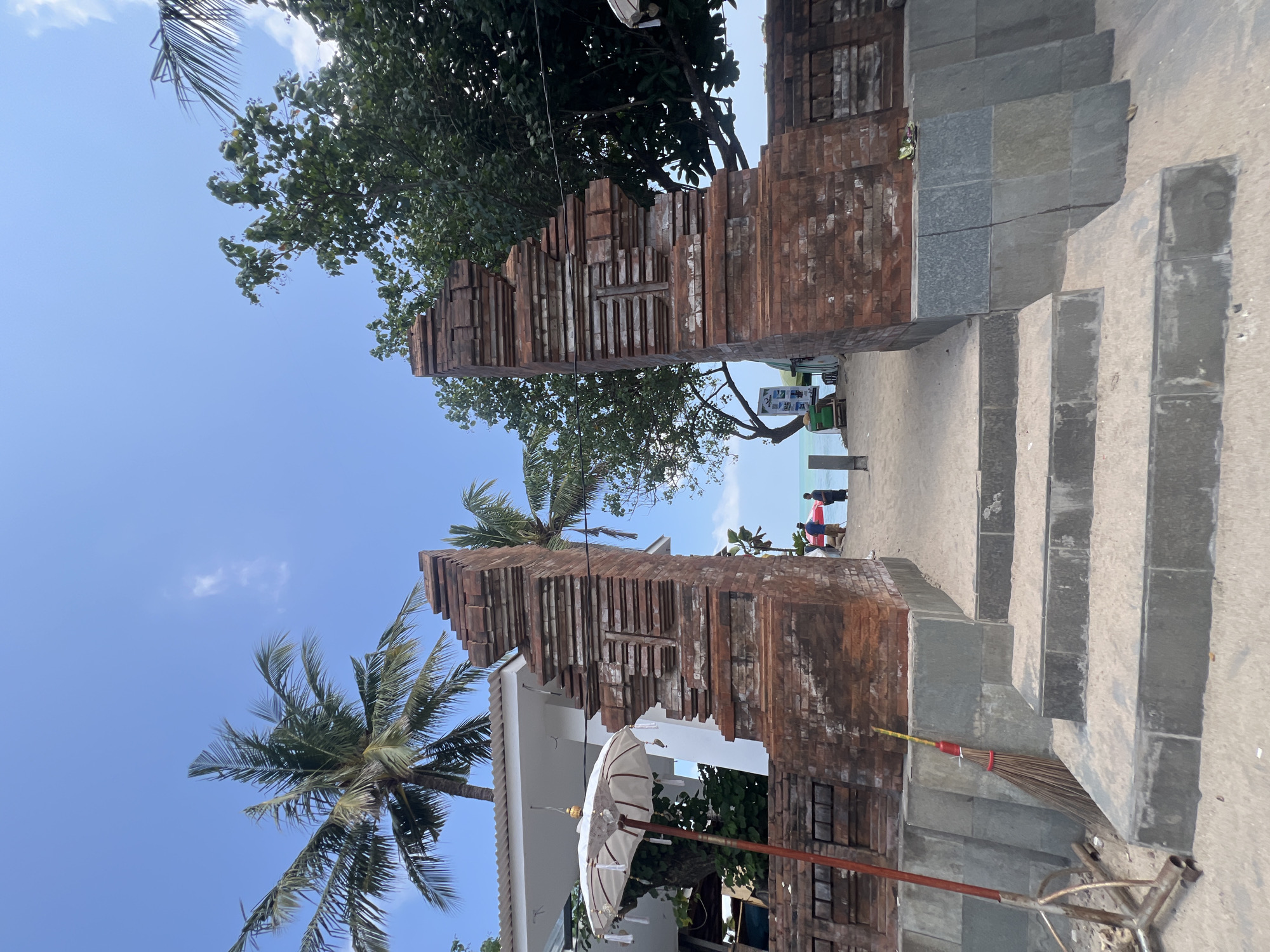 An outdoor, ground-level, eye-level, medium shot captures a weathered brick archway in Kuta, Indonesia, leading to a sandy beach and the ocean. The archway, constructed of red bricks with a tiered, textured design, is flanked by palm trees and lush green foliage. To the left of the arch, a white umbrella with tassels stands beside a white building with a white roof. A broom leans against the base of the archway. In the distance, on the beach, two figures stand near a red umbrella, and the ocean meets the horizon under a clear blue sky with a few wispy clouds. The foreground shows a set of stone steps leading up to the archway, with sand on either side.