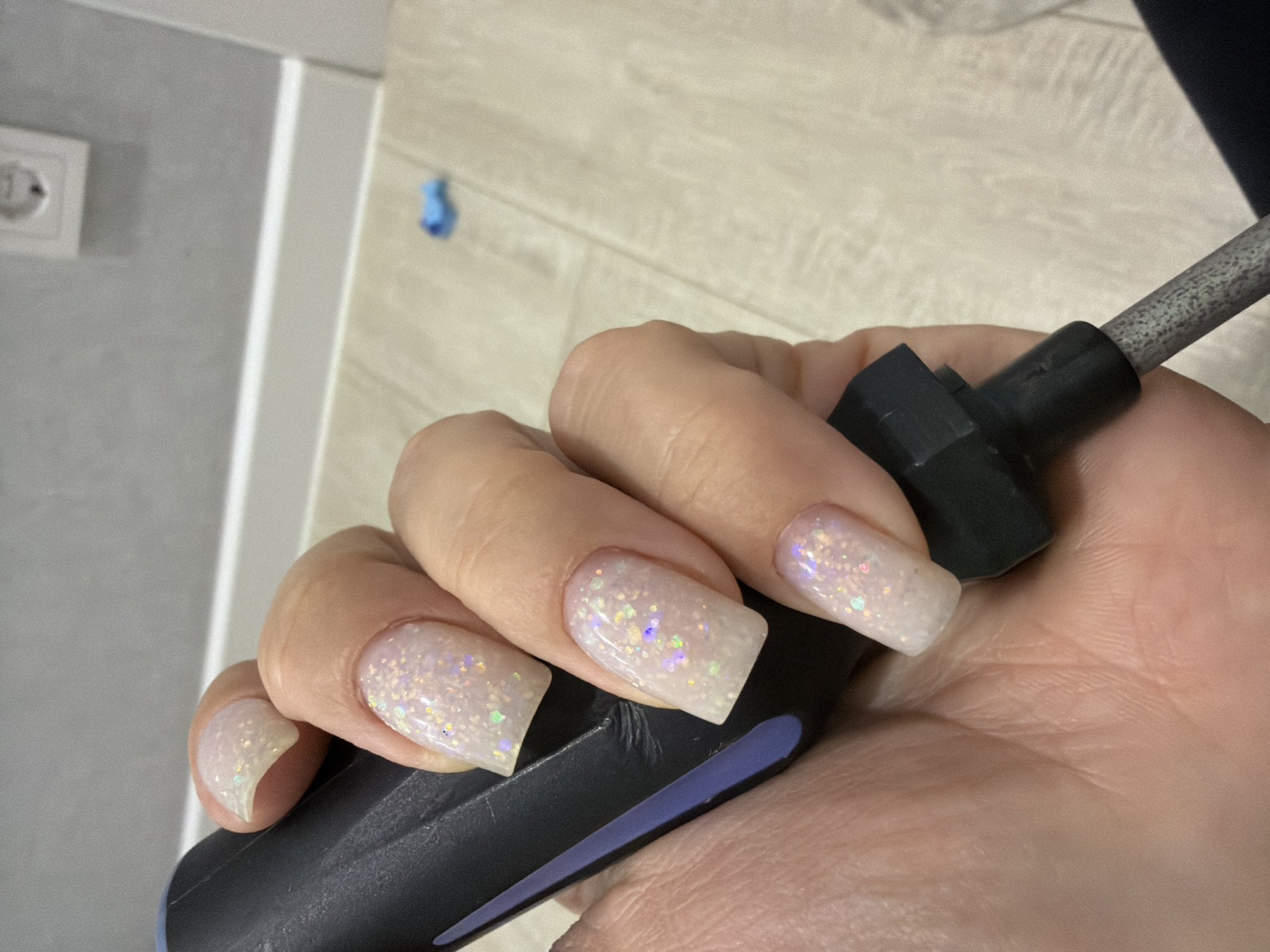 A close-up shot depicts a fair-skinned hand holding a black and grey metal tool with a plastic handle. The hand features long, square-shaped fingernails with a pale, glittery polish. The background includes a light-colored wood-look flooring and a white wall with a light switch and a blue object attached. The location, Votkinsk, Russia, cannot be directly discerned from the visual information provided in the image.