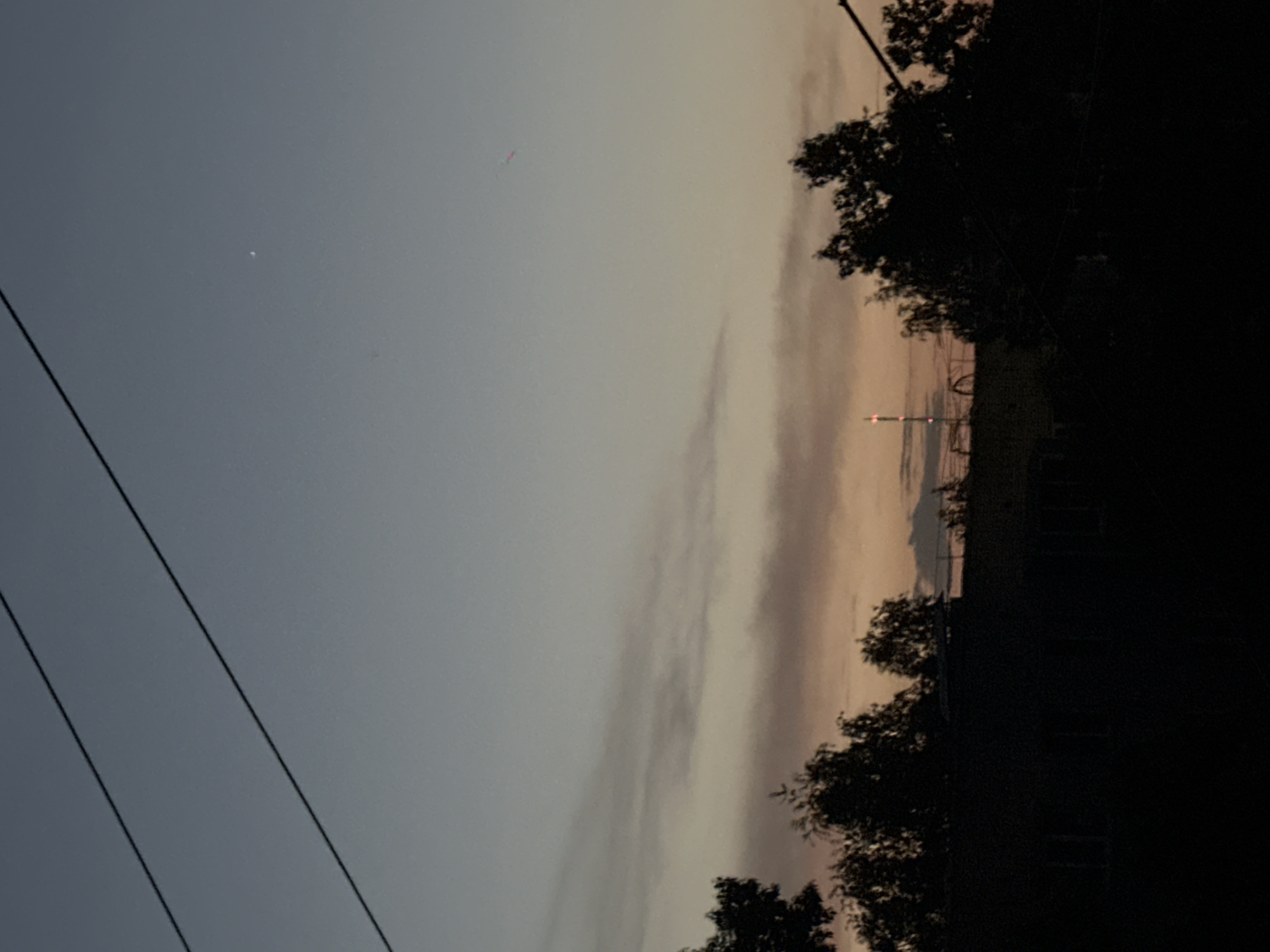 The media file captures a twilight sky over Biysk, Russia. The scene is dominated by a vast expanse of sky transitioning from a deep blue to lighter shades of gray and pale orange, indicating either dawn or dusk. wispy clouds are visible, adding texture to the sky. In the foreground, silhouetted against the sky, are the dark shapes of trees and power lines, suggesting an outdoor or semi-rural setting. In the distance, a tall structure, possibly a radio tower or a building, is visible with illuminated lights. The overall mood is serene and tranquil, typical of the quiet hours of early morning or late evening. There is no visible text in the image.