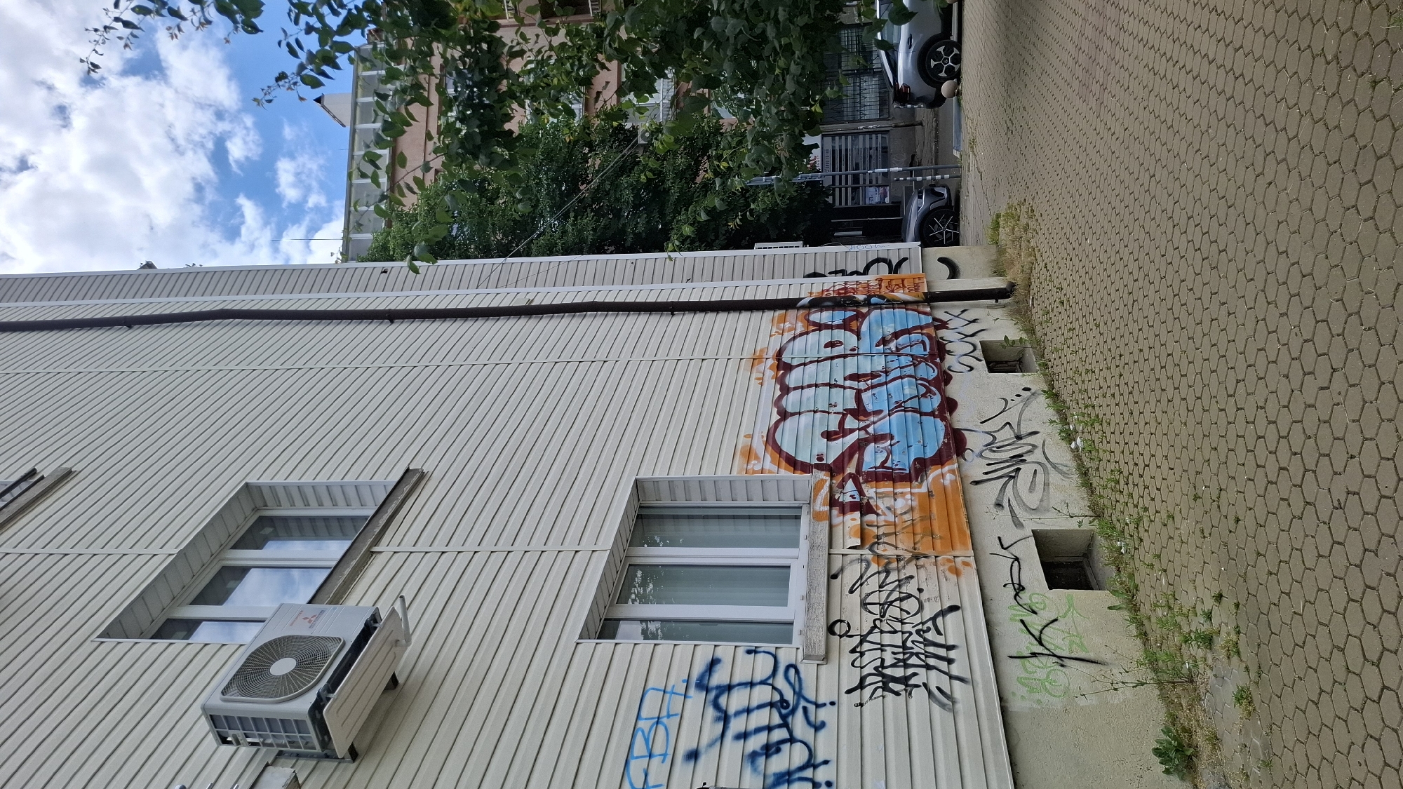 The image depicts the exterior of a building, likely residential, with light-colored siding. An air conditioning unit is mounted on the left side of the building, above a window. The lower portion of the building's wall is covered in graffiti, featuring colorful spray paint tags. A downspout runs vertically along the side of the building. The ground in front of the building is paved with hexagonal interlocking bricks. Vegetation, including grass and weeds, is growing along the base of the building and in patches on the sidewalk. In the background, trees and other buildings are visible, as well as two cars parked on a street. The sky is partly cloudy with blue patches. The style of graffiti and the urban setting suggest this location is in Sofia, Bulgaria.