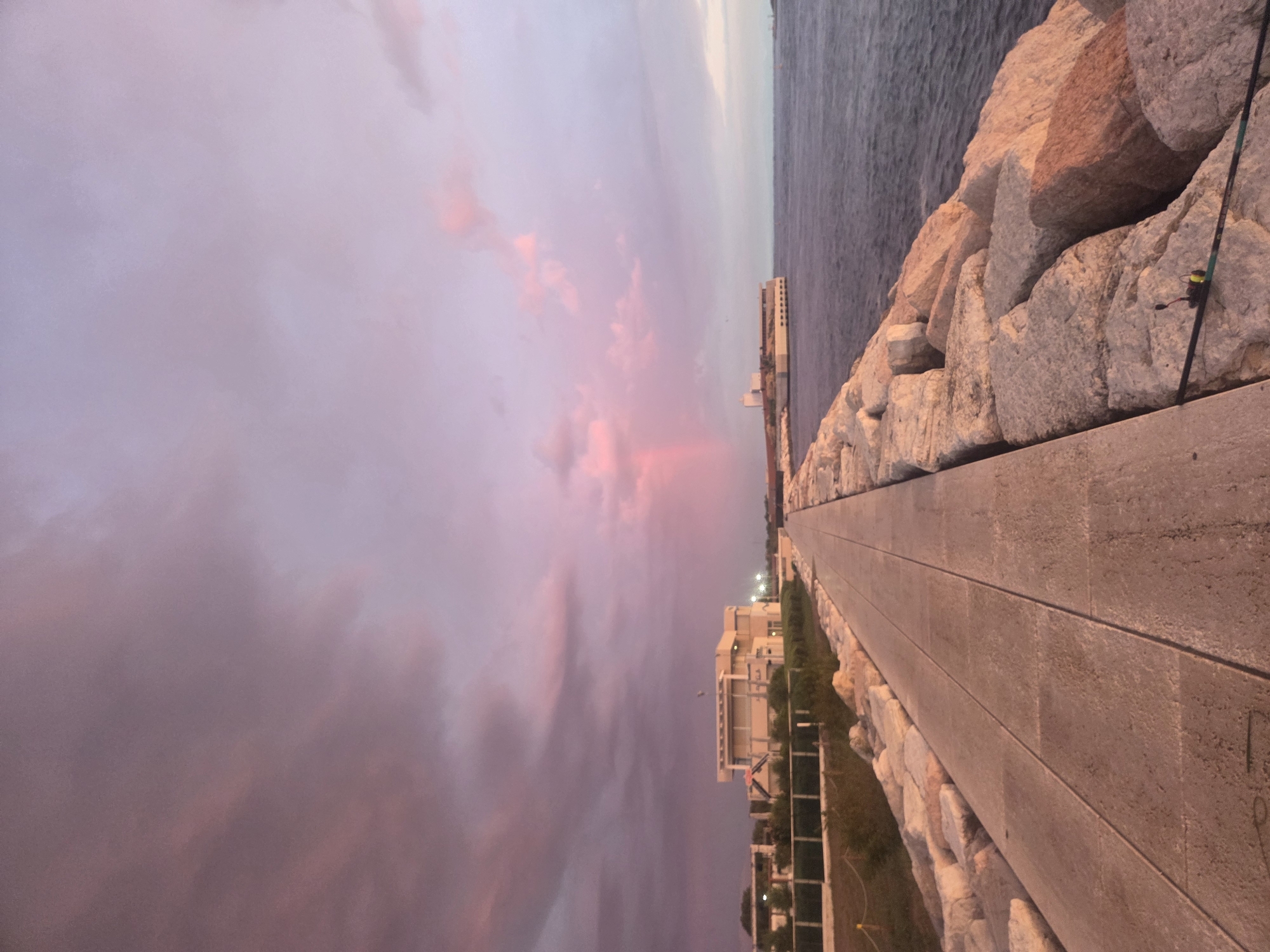 The image shows a paved walkway lined with large rocks on one side and a body of water on the other. In the distance, there are buildings and a faint rainbow in the sky. The sky is filled with pink and purple clouds, suggesting it is either sunrise or sunset. The scene appears to be a coastal area. Based on the architecture and coastal setting, this could be the Lungomare Adriatico in Chioggia, Italy.