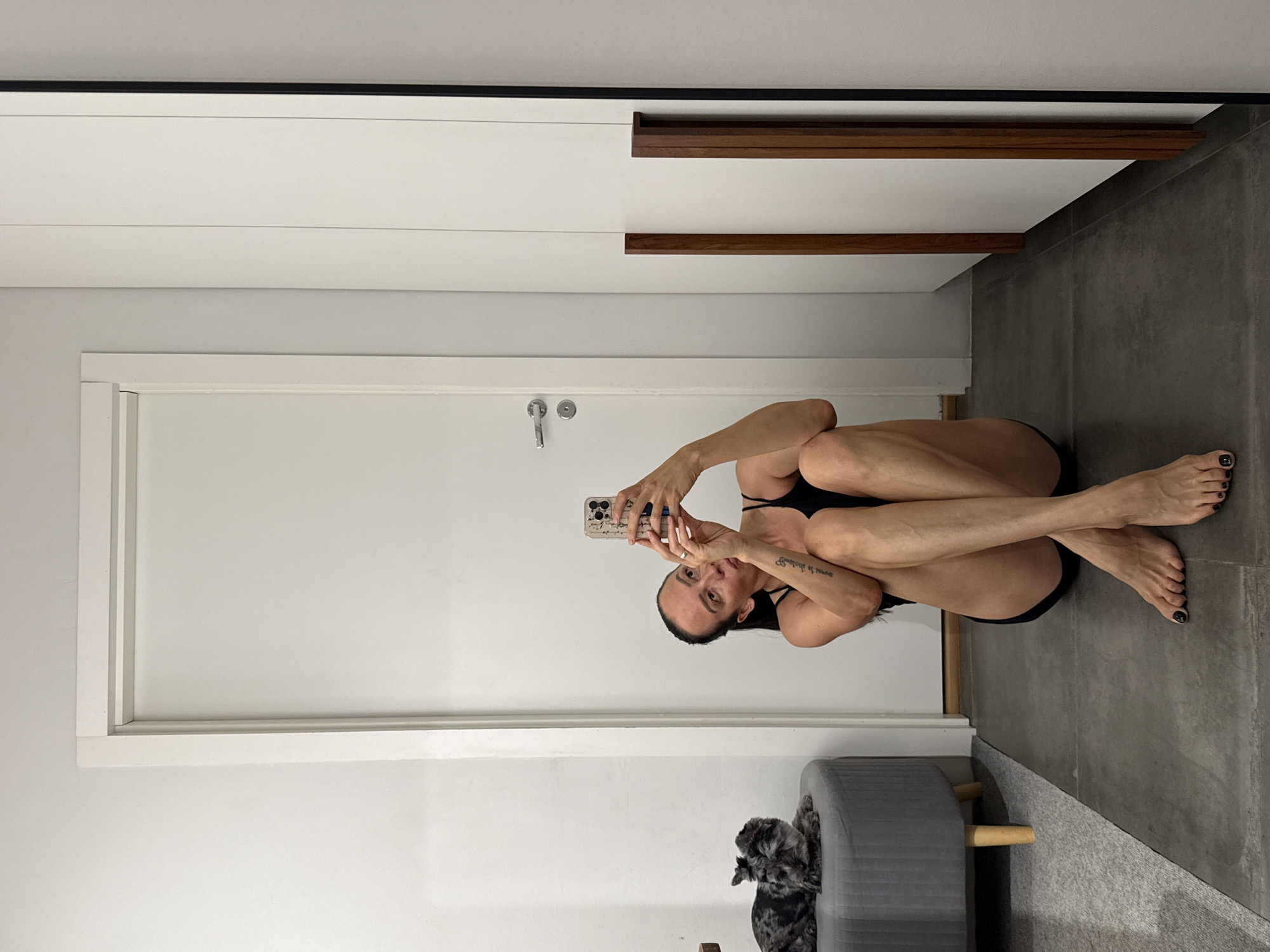 An adult female with dark hair pulled back in a ponytail is sitting on a dark gray tiled floor, facing a wall with a white door to her left and a white closet to her right. She is wearing a black tank top and black bottoms, and her legs are crossed. She is holding a smartphone in her right hand, taking a selfie. Her left arm is bent, resting on her left leg. A small, gray dog with dark spots is lying on a round, gray ottoman to her left. The environment appears to be an indoor residential space, likely a bedroom or hallway, with white walls and minimalist furnishings. The image is taken from a slightly low angle, looking up at the woman.