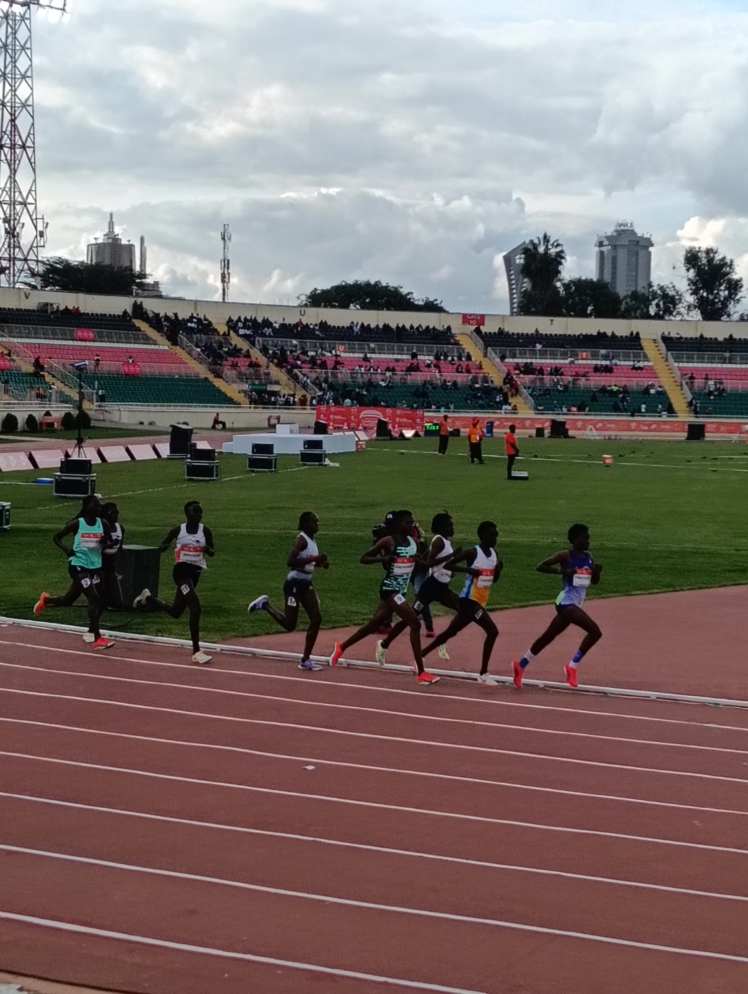 An outdoor long-distance running race is captured from a slightly elevated perspective within an athletics stadium in Nairobi, Kenya. Seven female runners, distinguished by their athletic wear and bib numbers, are actively competing on the red synthetic track in the foreground. Their focused expressions and running forms suggest a competitive moment during the event.
The stadium features multi-tiered seating, with some sections occupied by spectators and others appearing empty. Green grass covers the infield, and various equipment like electronic displays and speaker systems are visible near the track. In the background, under a heavily cloudy sky, tall communication towers and city buildings are discernible, indicating an urban setting.
Notable details include a "GATE 10" sign visible on a yellow section of the stands. Several runners have partially legible names or sponsors on their vests, such as "EMELIA," "KIPSENG," "CHEMENG," and "KIPSANG," along with bib numbers like "8," "19," and "24." A digital clock or timer on the infield shows "7:3X," possibly indicating elapsed race time. The overall lighting suggests it is either late afternoon or early evening, given the diffused light and overcast conditions. A few stadium staff or officials in orange vests are also present on the infield.