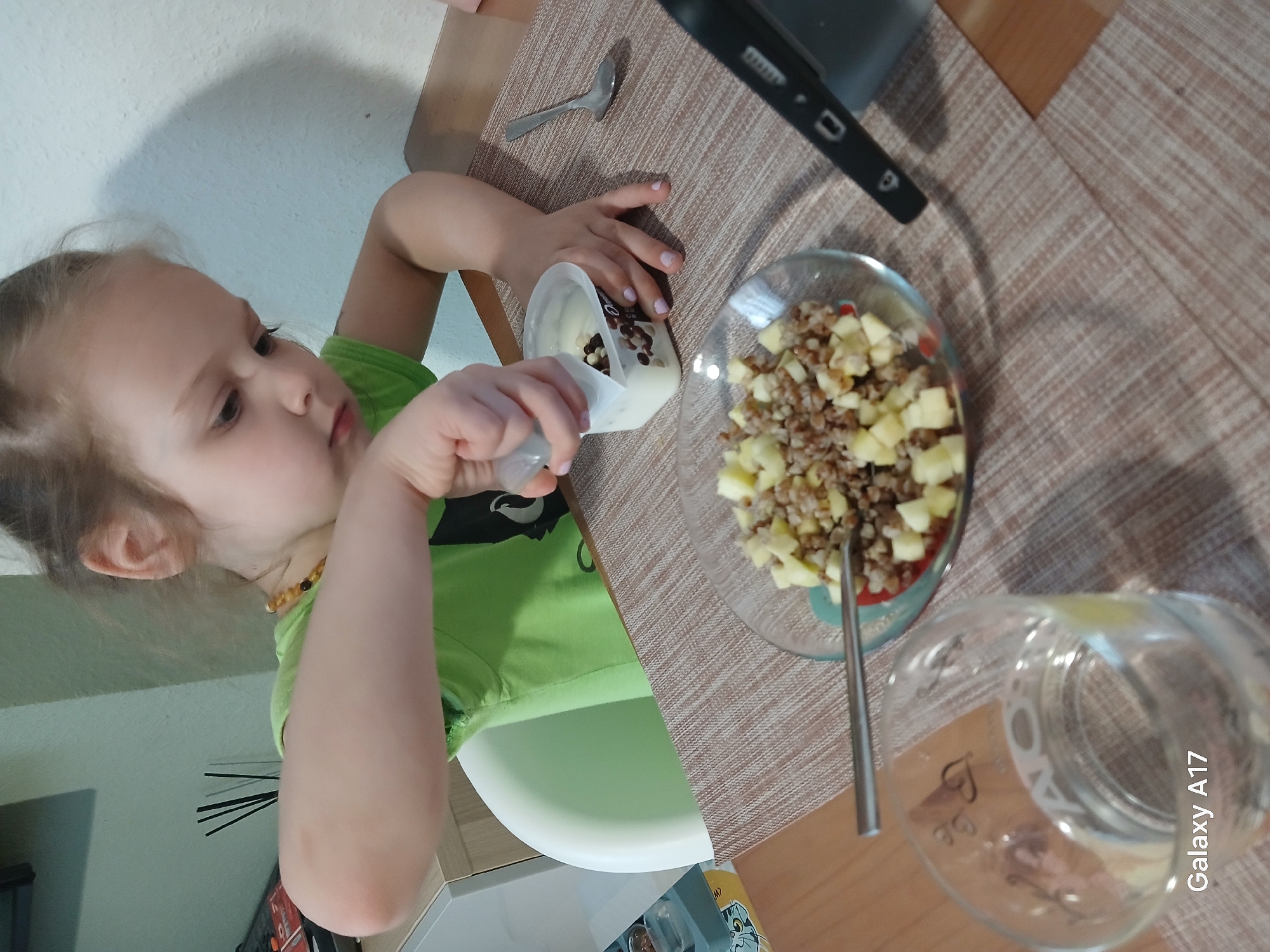 A young child in a green shirt is pouring chocolate chips from a white container into a salad in a glass bowl on a brown textured tabletop. A spoon and a glass of water are also on the table. A smartphone and a silver spoon are nearby. The image was taken in Yekaterinburg, Russia.