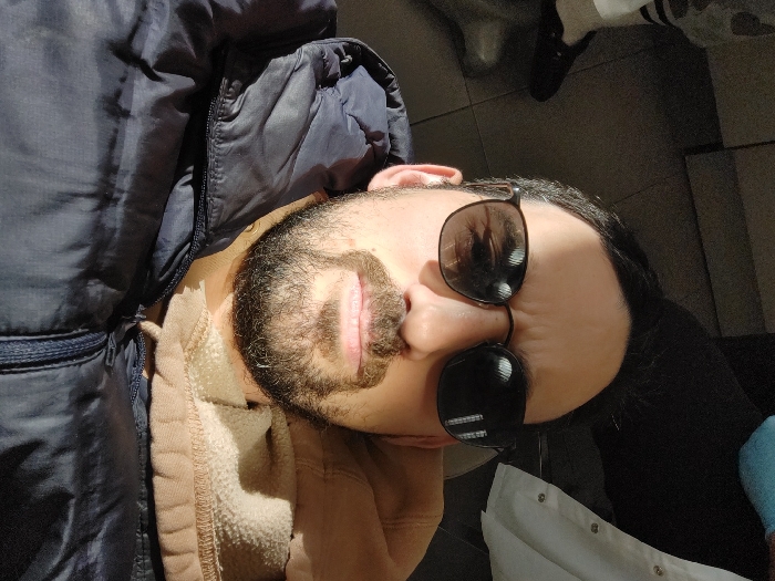 A male individual is depicted in a close-up, upward-angled selfie photograph taken in an indoor setting in Tel-Aviv, Israel. The male subject has dark hair, a beard, and a mustache. He is wearing dark-framed sunglasses with dark, reflective lenses. His attire consists of a dark blue, possibly puffer-style, zipped jacket worn over a tan-colored hooded sweatshirt, the hood of which is visible around his neck. A bright light source illuminates the upper left portion of his face and forehead, casting a shadow across his right eye. In the blurred background, to the upper left, the lower leg and foot of a second individual are partially visible, wearing light-colored pants with dark stripes and a dark slip-on shoe. To the upper right background, a segment of a white garment, resembling a lab coat or uniform with visible buttons, is present, alongside a portion of light blue fabric or object. The floor, visible in the upper left, is dark and tiled.