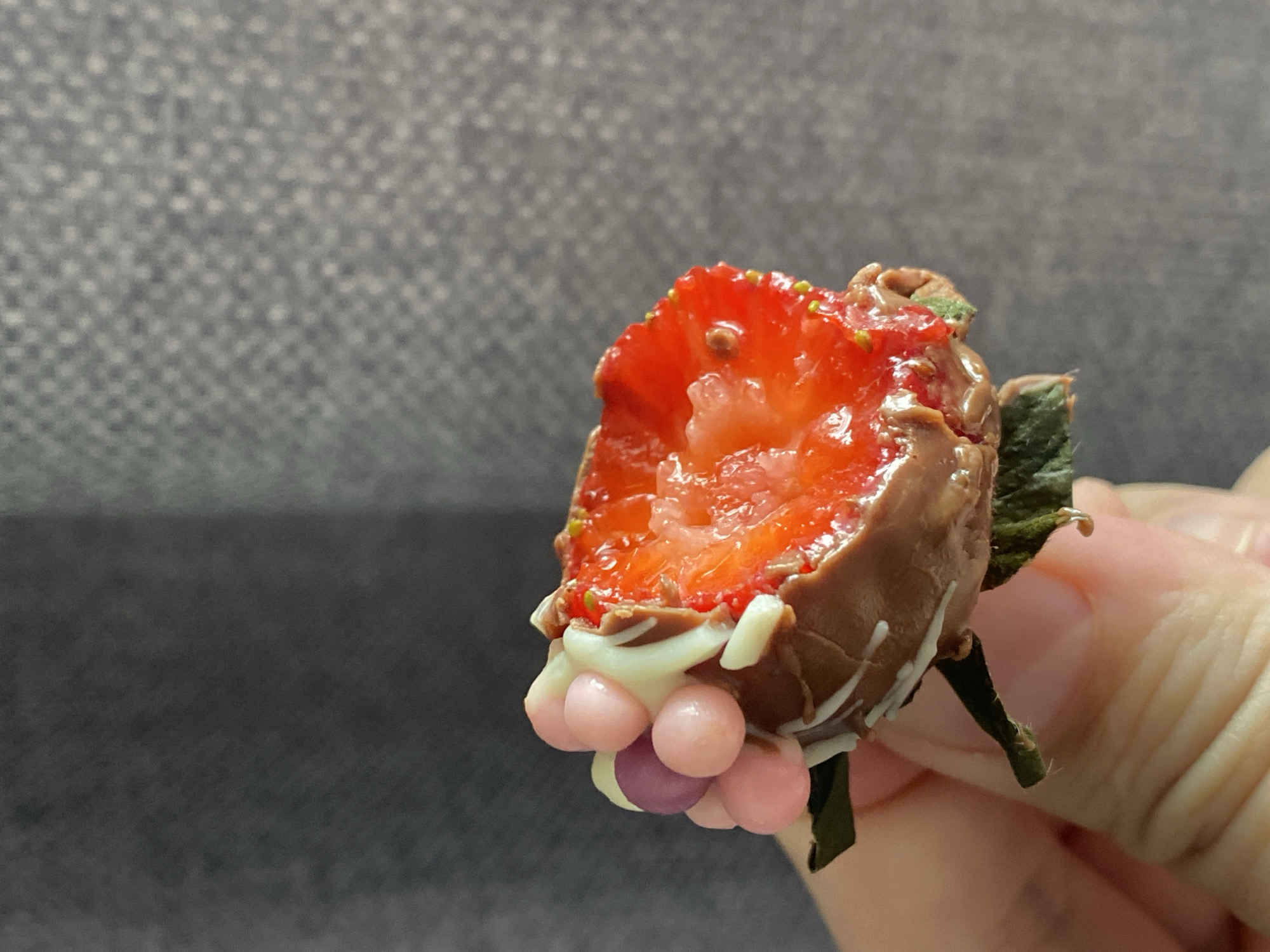 A person's hand holds a chocolate-covered strawberry with a portion cut out and decorated with pink and purple candy balls, and white drizzle. The background is blurred and appears to be a gray fabric surface. The city is Unknown, Unknown.