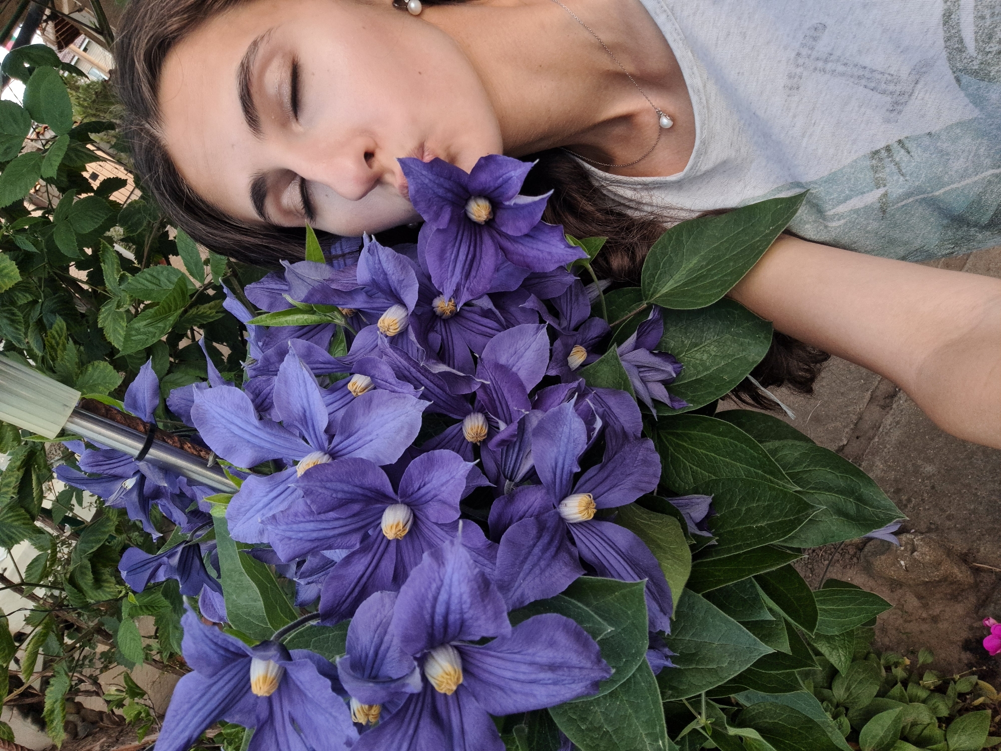 A young woman with brown hair lies among a profusion of purple clematis flowers with green leaves. Her eyes are closed, and she appears to be touching or kissing the flowers with her lips. She wears a light-colored T-shirt and a delicate silver necklace with a pearl pendant. The background shows a mix of foliage and what appears to be a stone or brick surface. The location appears to be outdoors, perhaps a garden setting in an unidentified city.